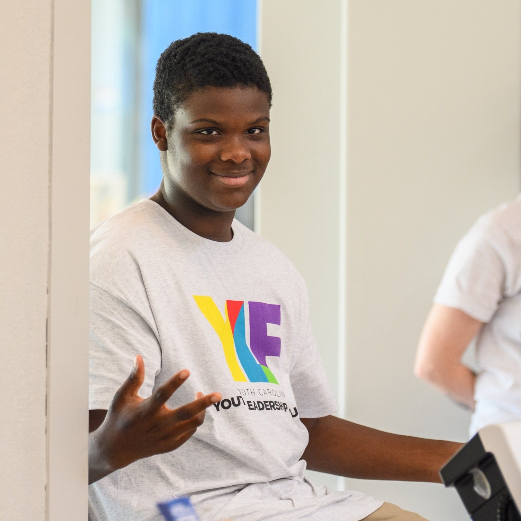 YLF delegate, a young Black man wearing the YLF t-shirt, smiles while seated and gestures to the camera.