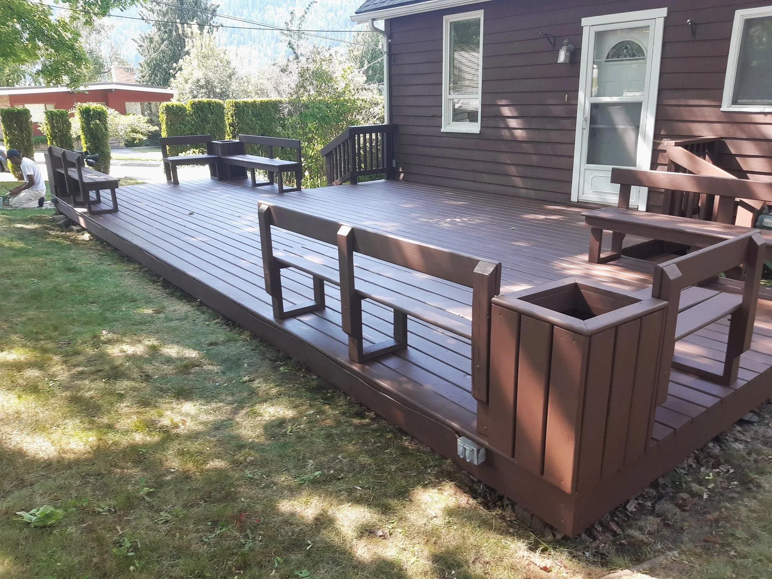 New wooden deck attached to a house with benches, planters, and a white door, surrounded by greenery and nearby houses.