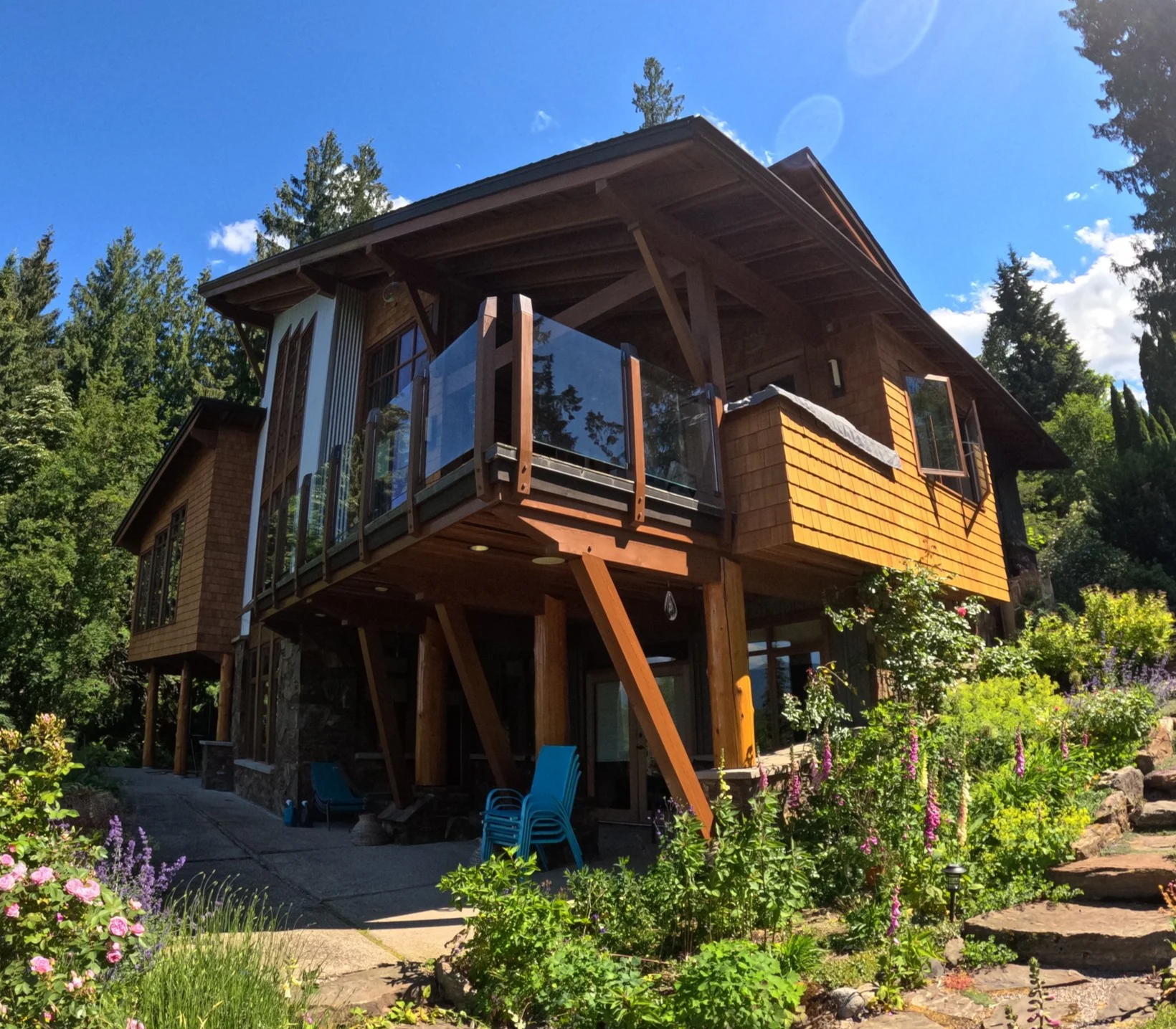 A modern house with a wooden exterior and large windows, elevated on stilts, set in a lush garden with flowers and trees, under a bright blue sky.