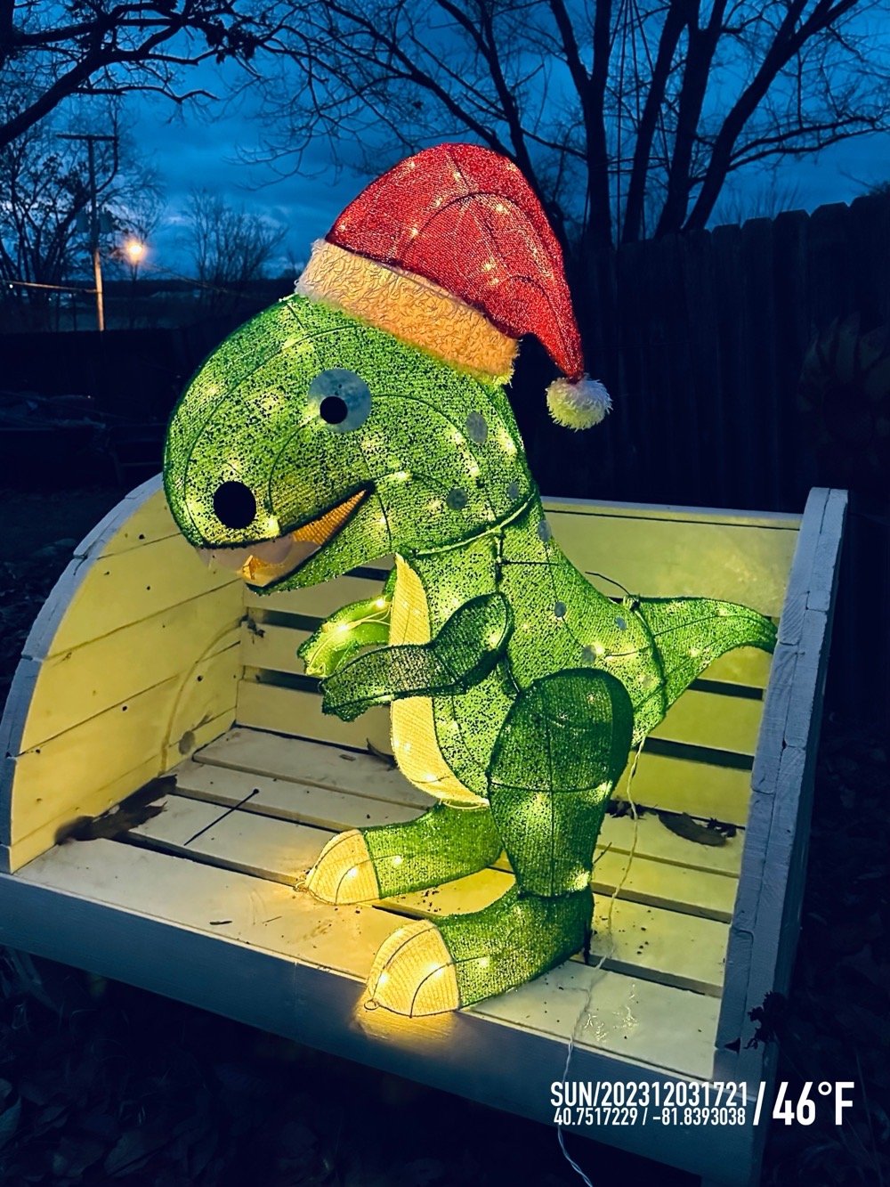 a green dinosaur in a santa hat with christmas lights for innards sitting on a yellow bench at dusk.