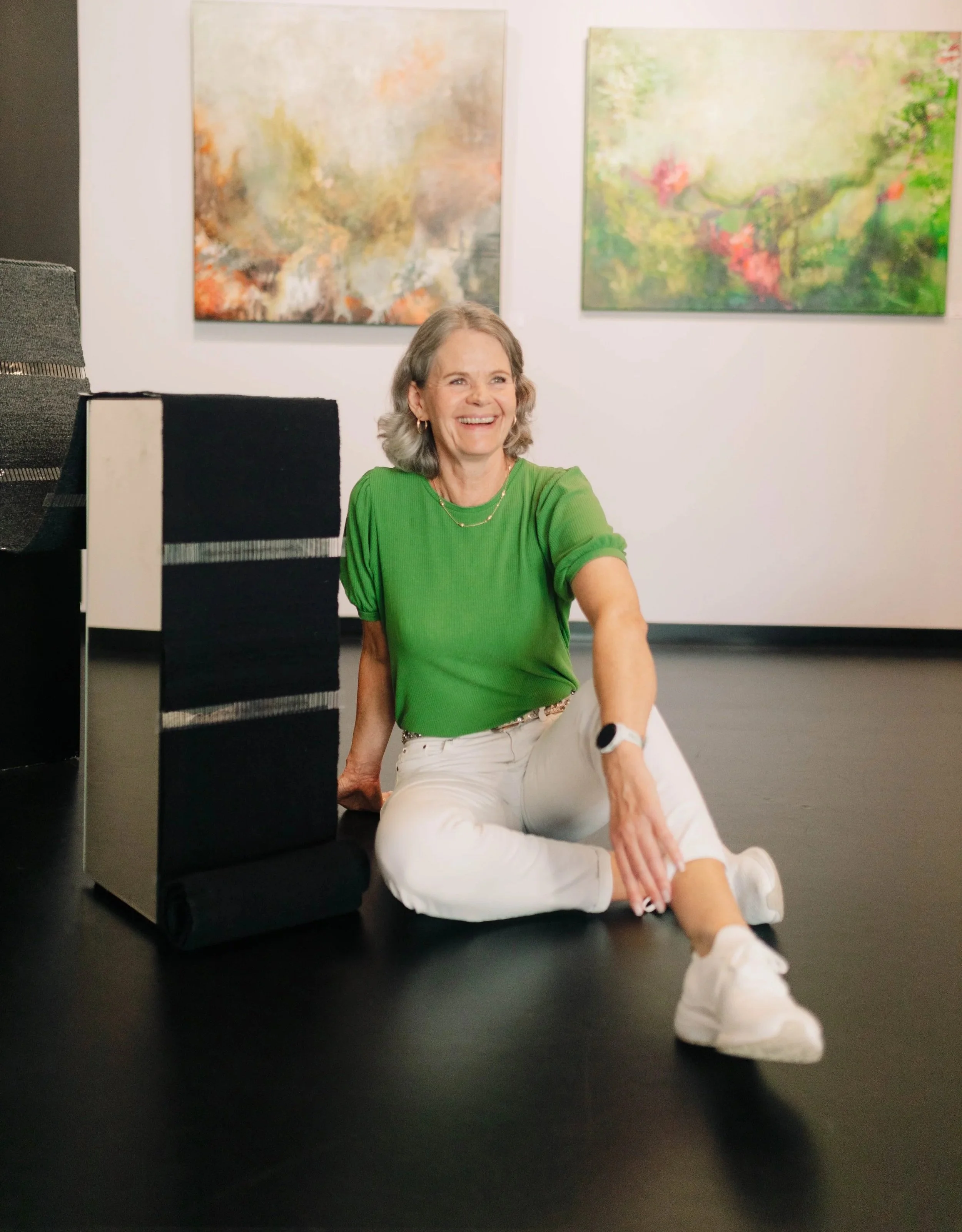 Gro Moe sitter på galverigulv foran abstrakte malerier under utstilling