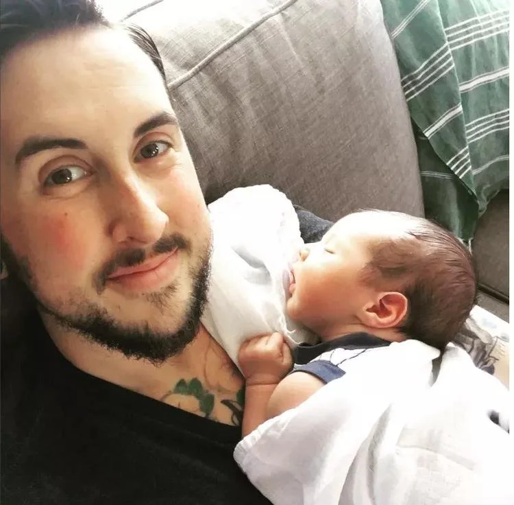 Joyful 30-something man with beard looks smiling at the camera while holding sleeping newborn baby