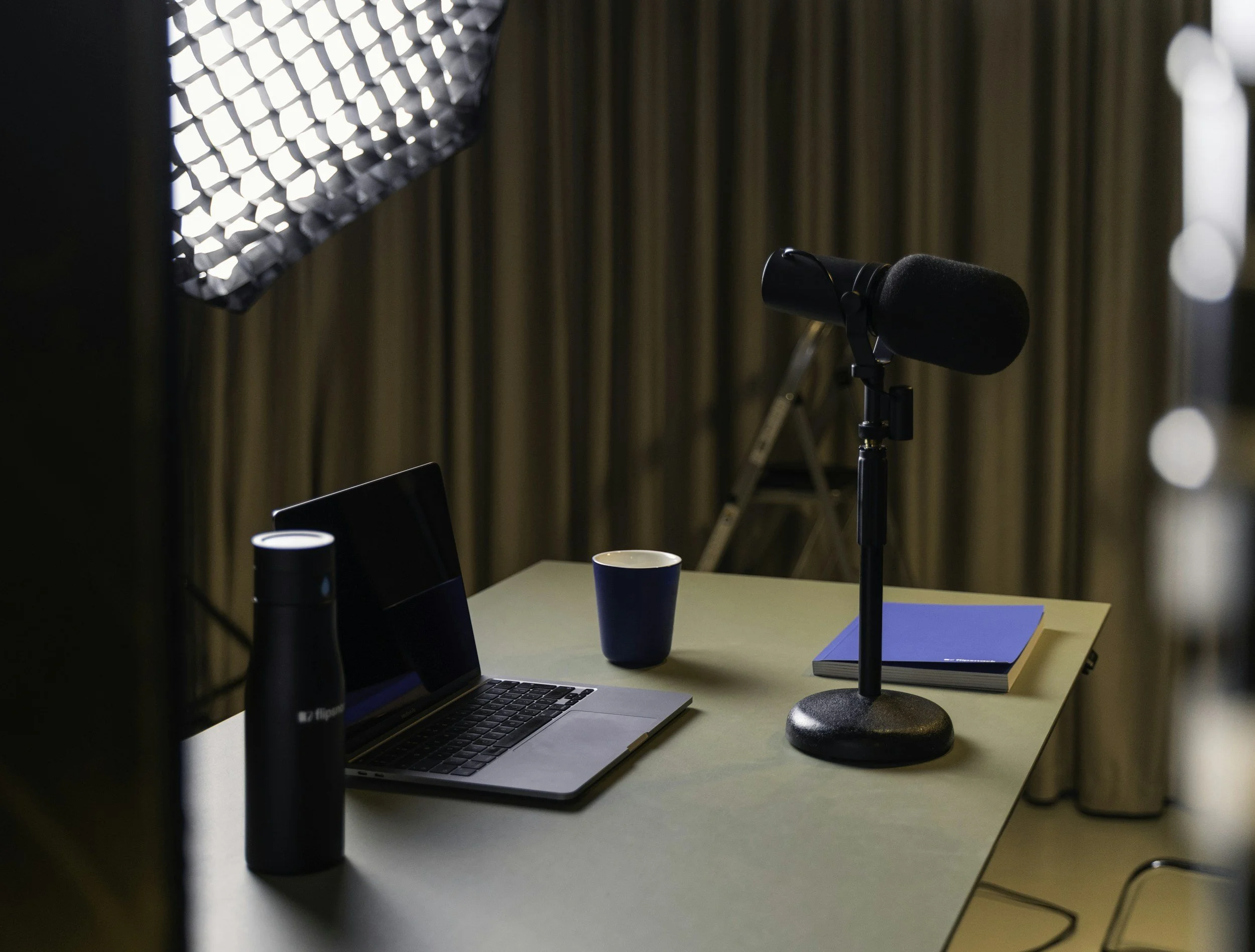 A workstation with a laptop, two black thermos bottles, a blue cup, a blue notebook, and a microphone on a desk. There are curtains in the background and a soft light source in the upper left corner.
