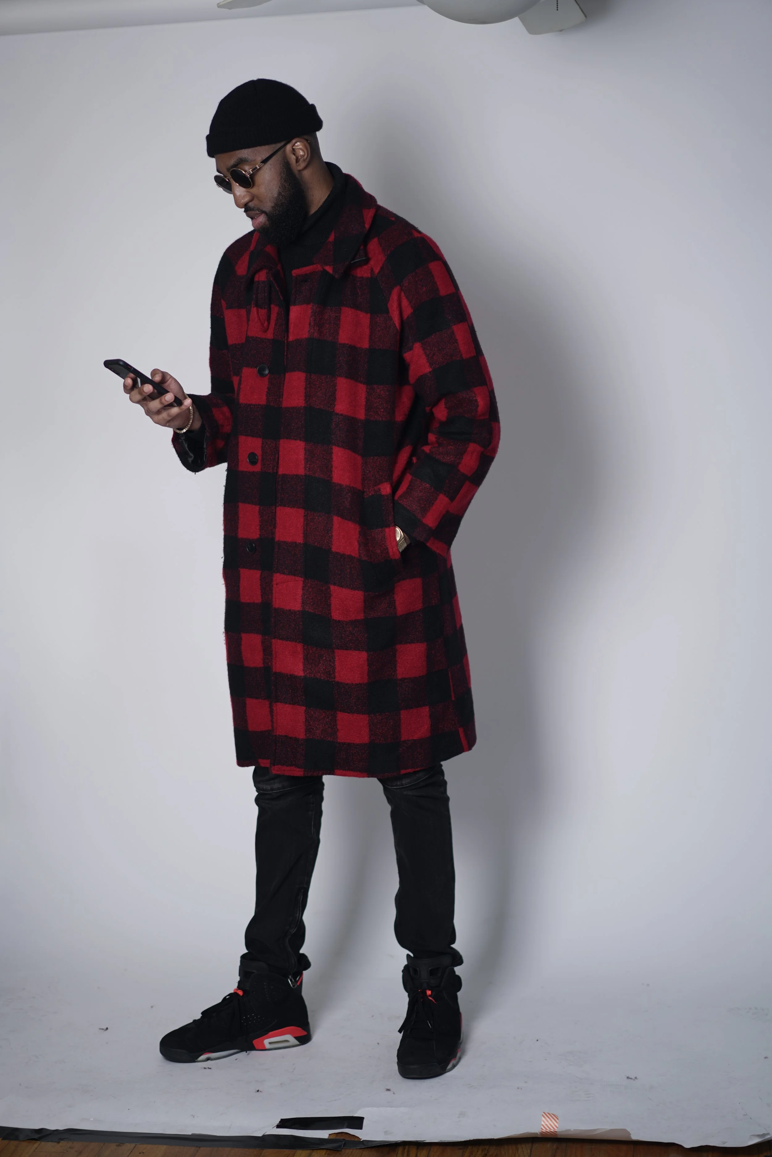 A man standing against a white background, wearing a black beanie, sunglasses, a black turtleneck, a red and black plaid coat, black pants, and black Nike sneakers with red accents, looking at his phone. Steven F. Thesier