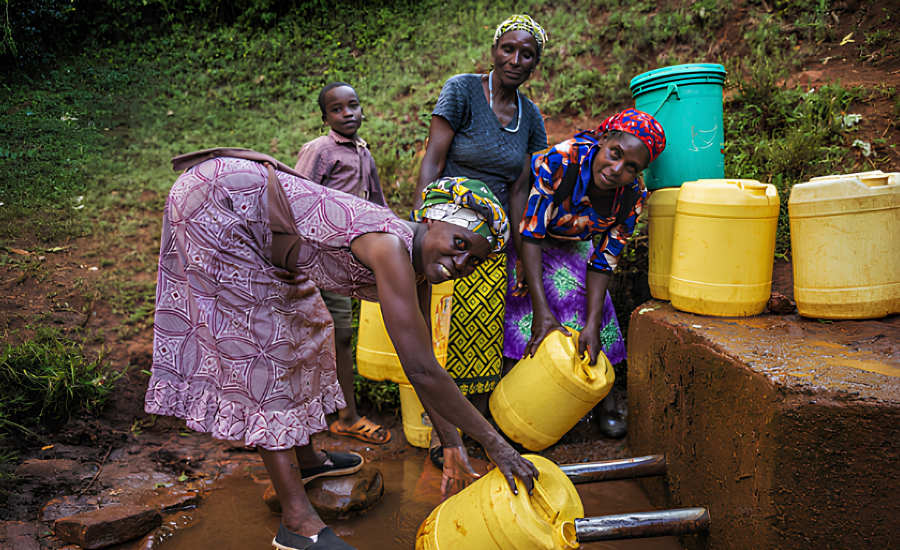 Women and a child gather water in yellow containers at an outdoor water source in a rural community.