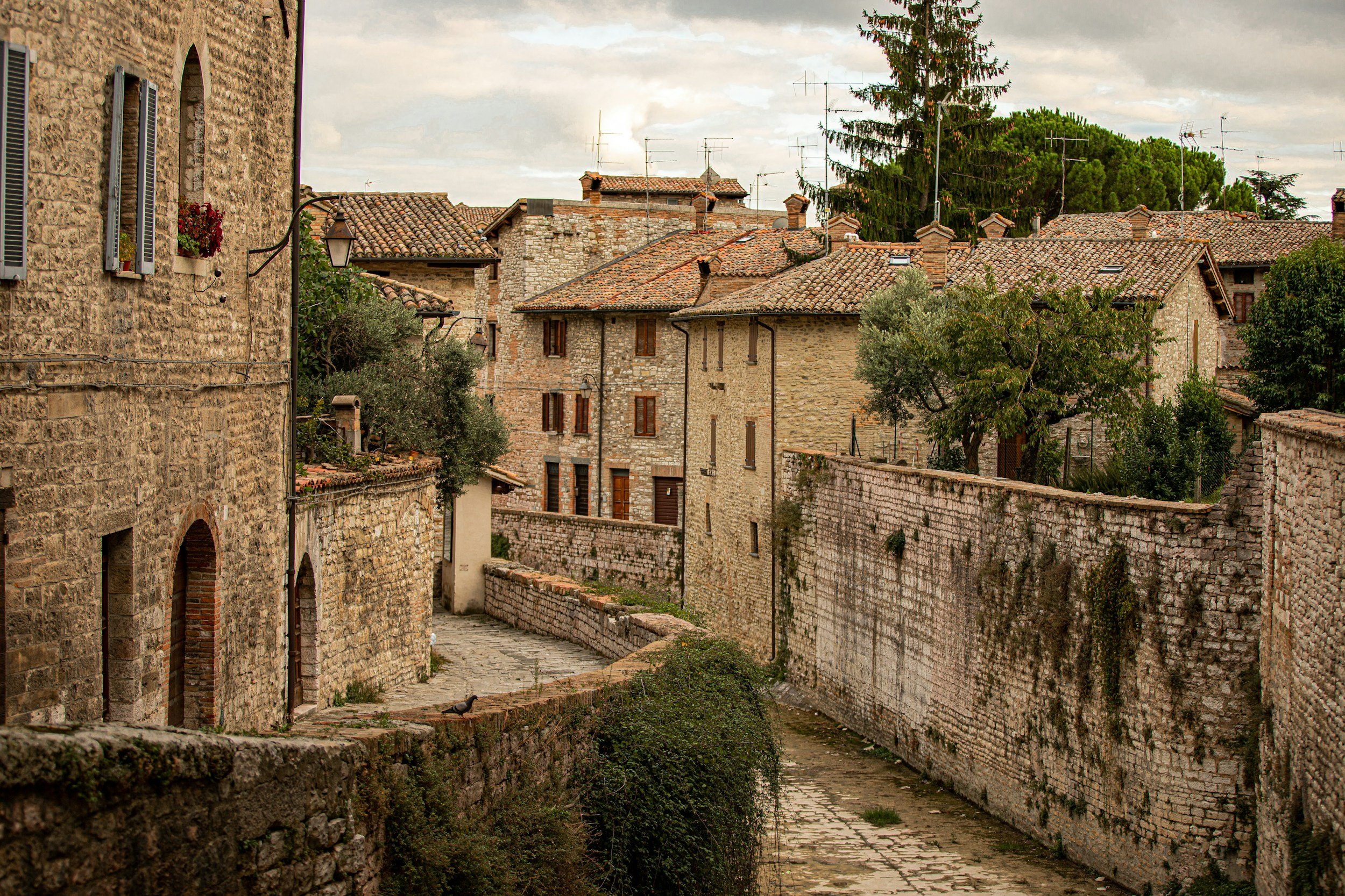 Historic stone buildings with tiled roofs, narrow pathway, and greenery, evoking a medieval European village atmosphere.