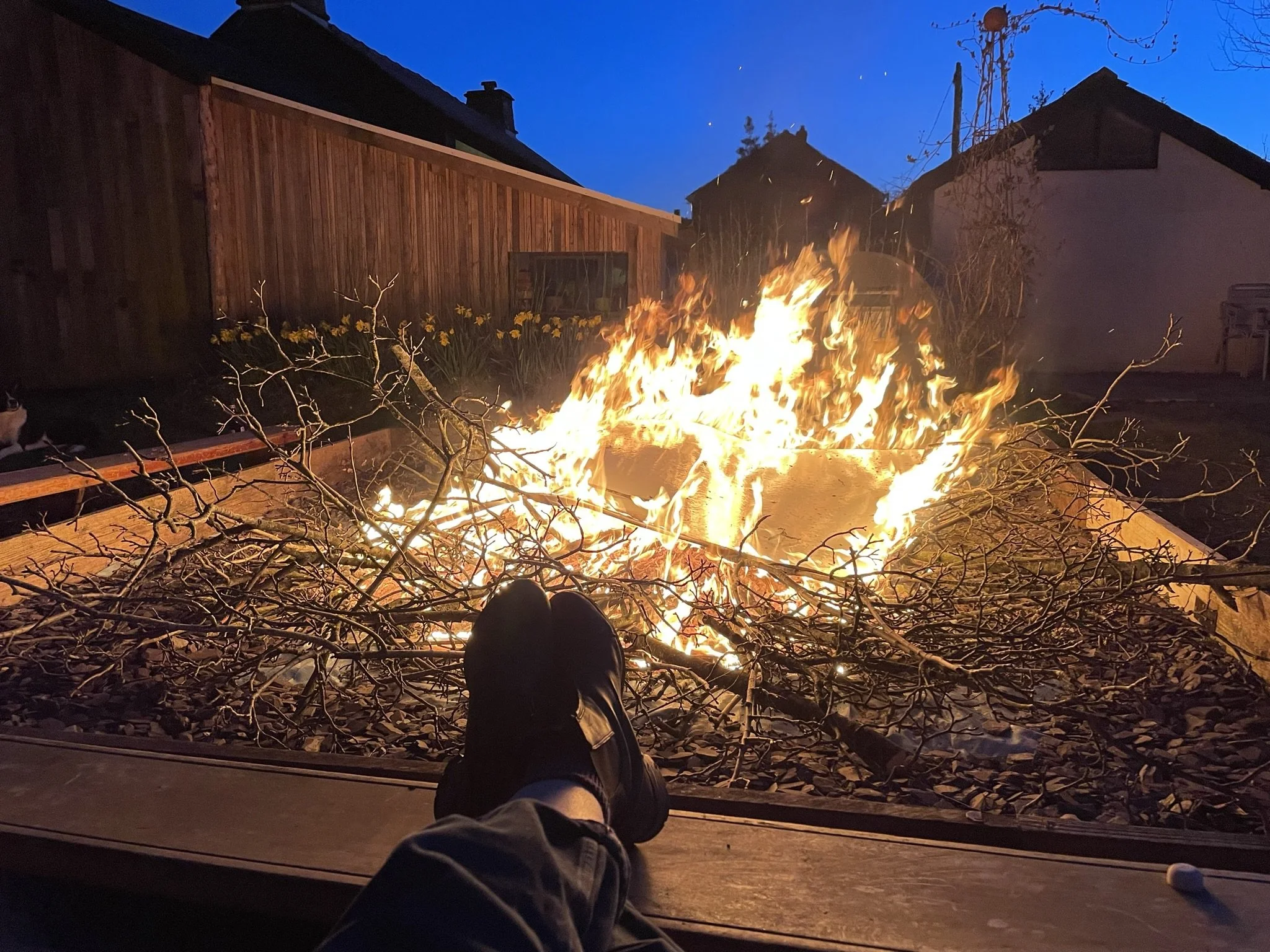 Person relaxing in backyard with feet up, watching a fire pit with burning branches at dusk.