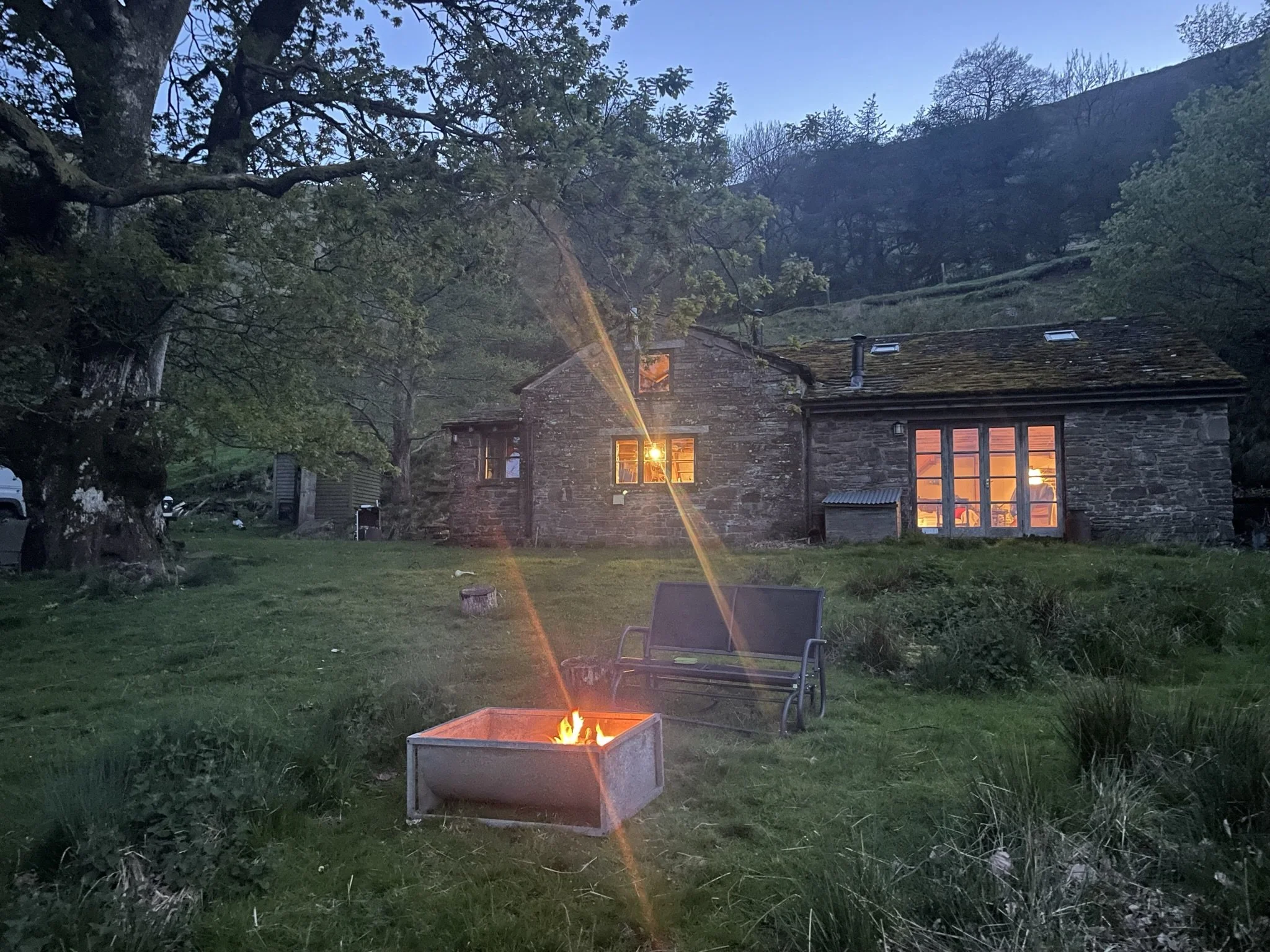 A stone house with lit windows at dusk, surrounded by trees and grass, with a fire pit and outdoor chairs in the foreground.
