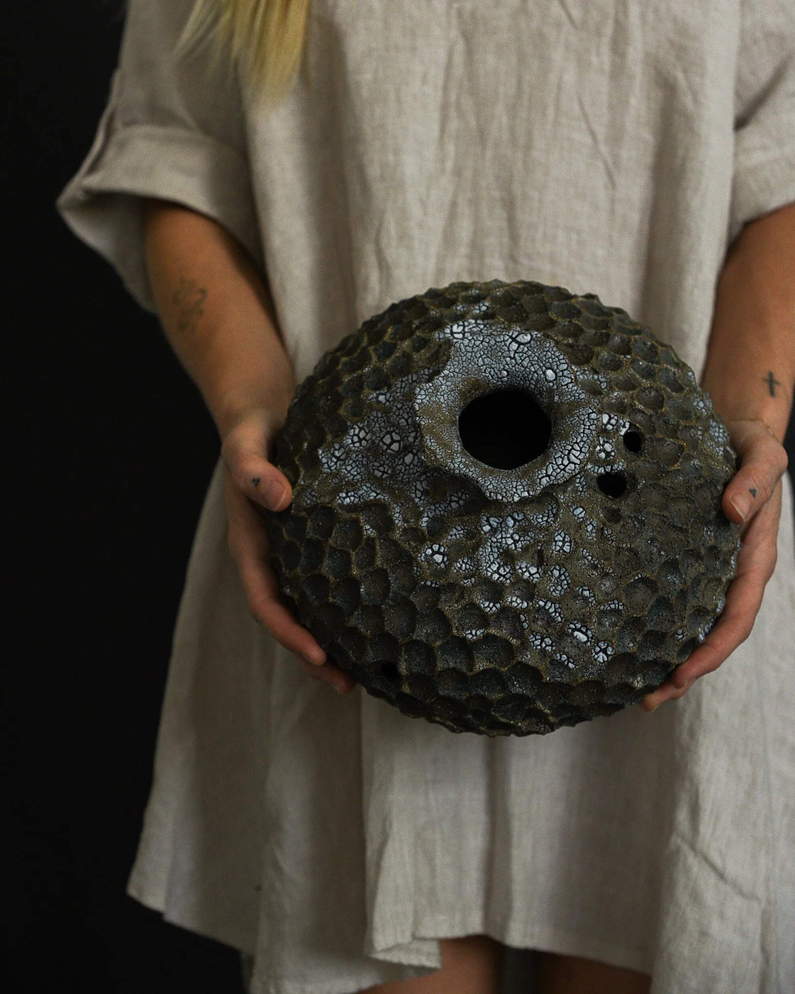 A person holding a hollow, textured, spherical object with a dark hole in the center. The person is wearing a beige or off-white shirt with part of a tattoo visible on their left arm.