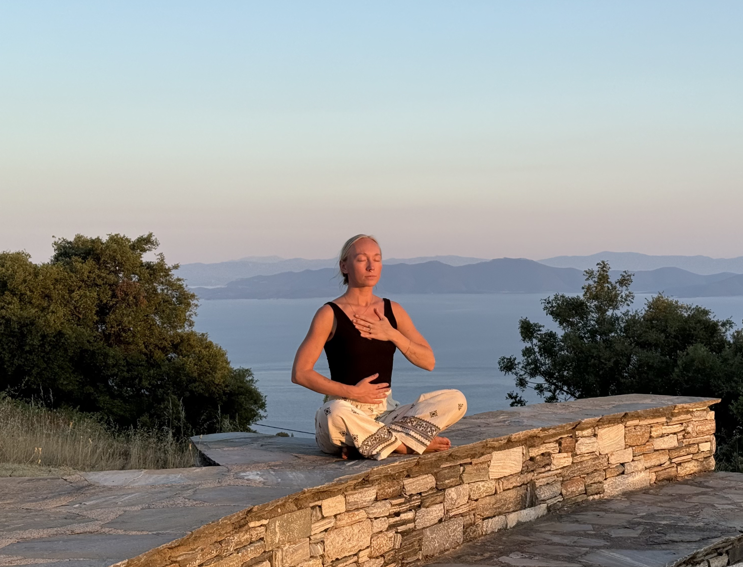 A woman practicing yoga outdoors on a stone platform, sitting cross-legged with her hands on her chest in a meditative pose, overlooking a lake and mountains at sunset.