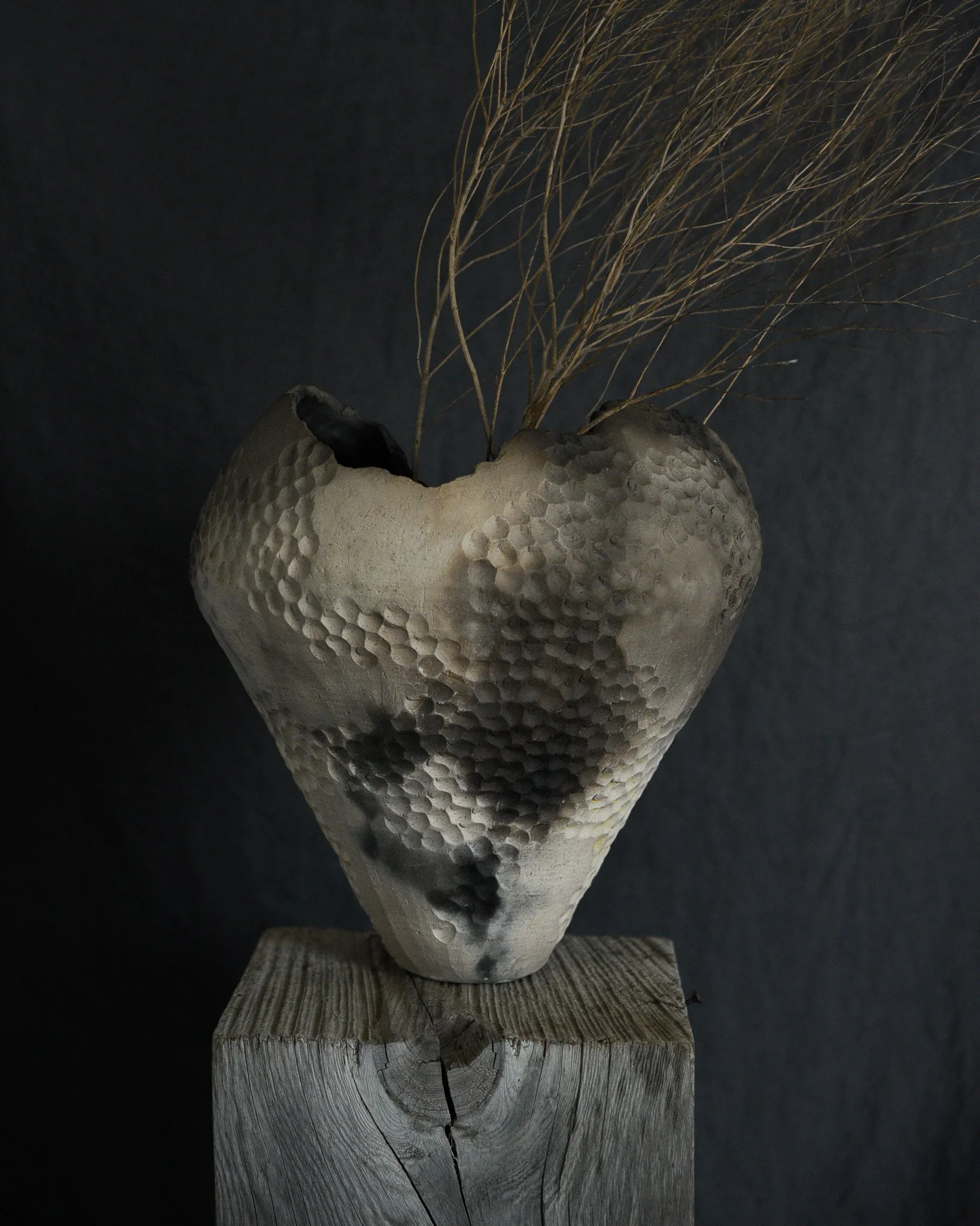 Ceramic vase with a textured surface and dried twigs, placed on a wooden block against a dark background.