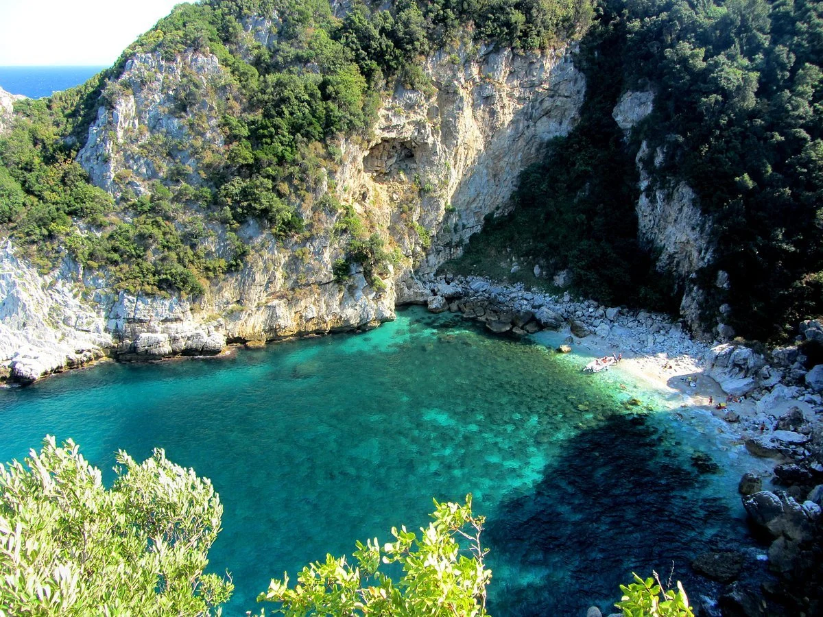 A secluded cove with turquoise water, surrounded by high rocky cliffs covered in lush green vegetation, and a small beach with a few people.