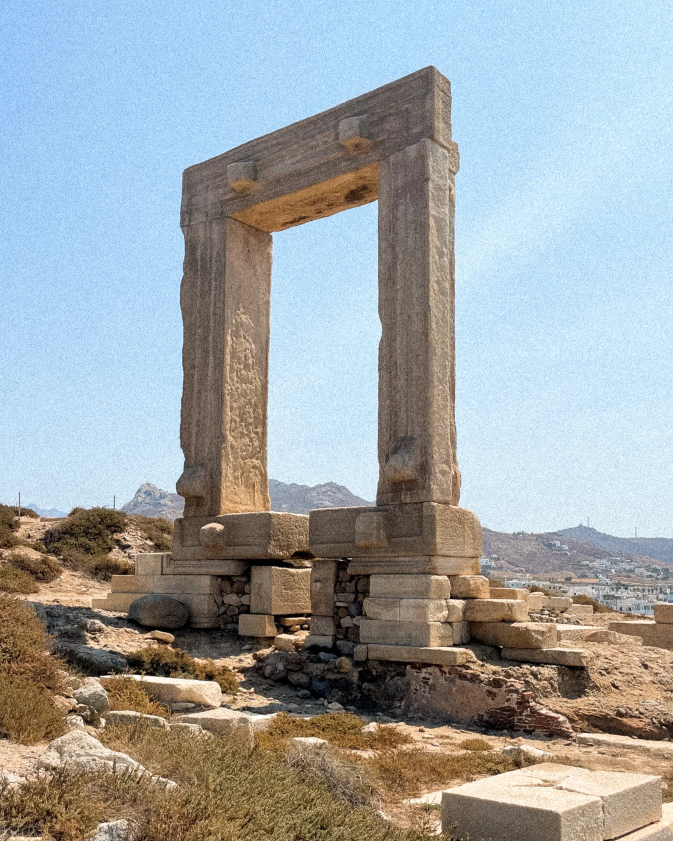 Ancient stone structure with two vertical columns supporting a horizontal lintel, situated in a desert landscape with mountains in the distance.