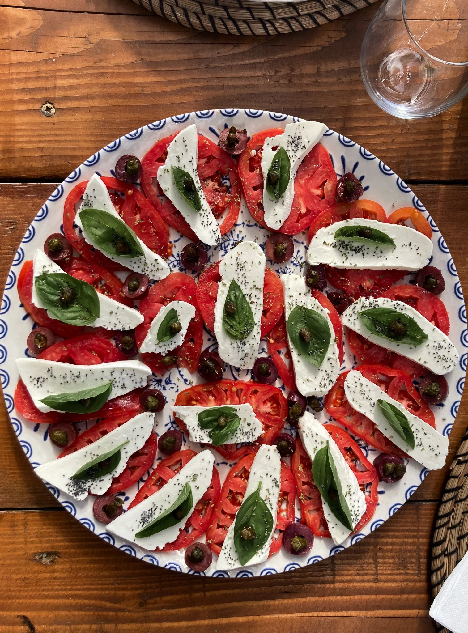 A plate with sliced tomatoes, slices of mozzarella cheese, fresh basil leaves, black pepper, and green olives, on a wooden table.