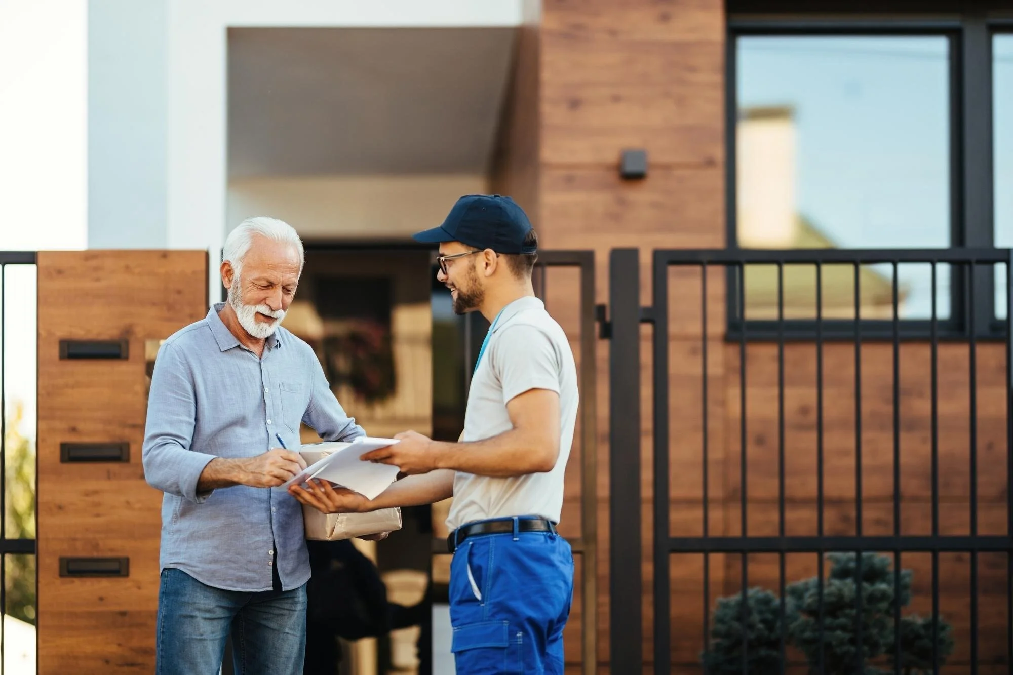 Two men are outside a house, one is holding a package, and they appear to be engaged in conversation