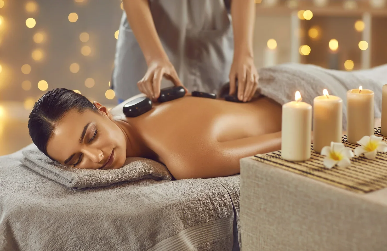 A woman receiving a hot stone massage at a spa, lying face down on a massage bed with candles and flowers nearby, creating a relaxing atmosphere.