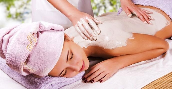 A woman lying on her stomach receiving a massage with white cream or lotion on her back, wearing a towel and a pink head towel.