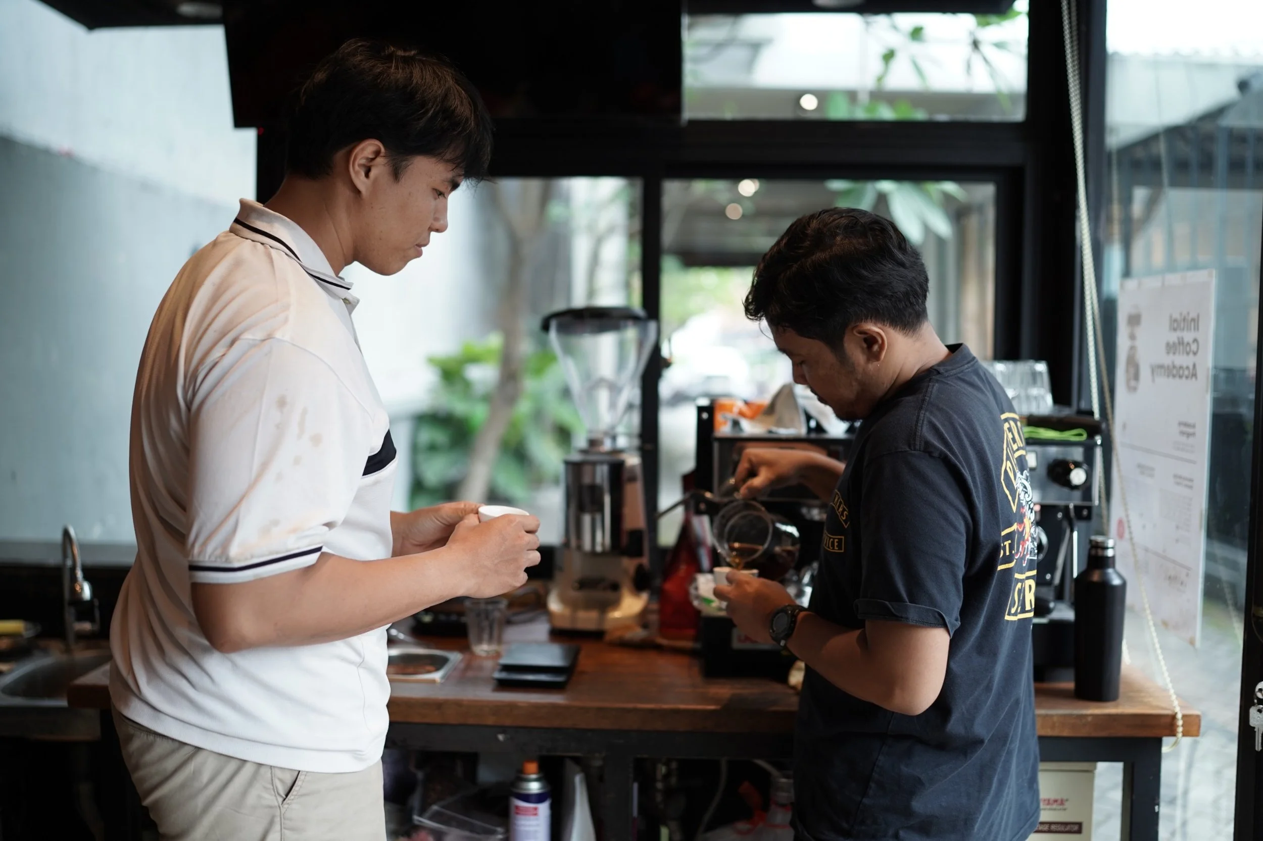 Two men in a coffee shop, one wearing a white shirt and the other wearing a black T-shirt, are standing at the counter and looking down at their drinks.