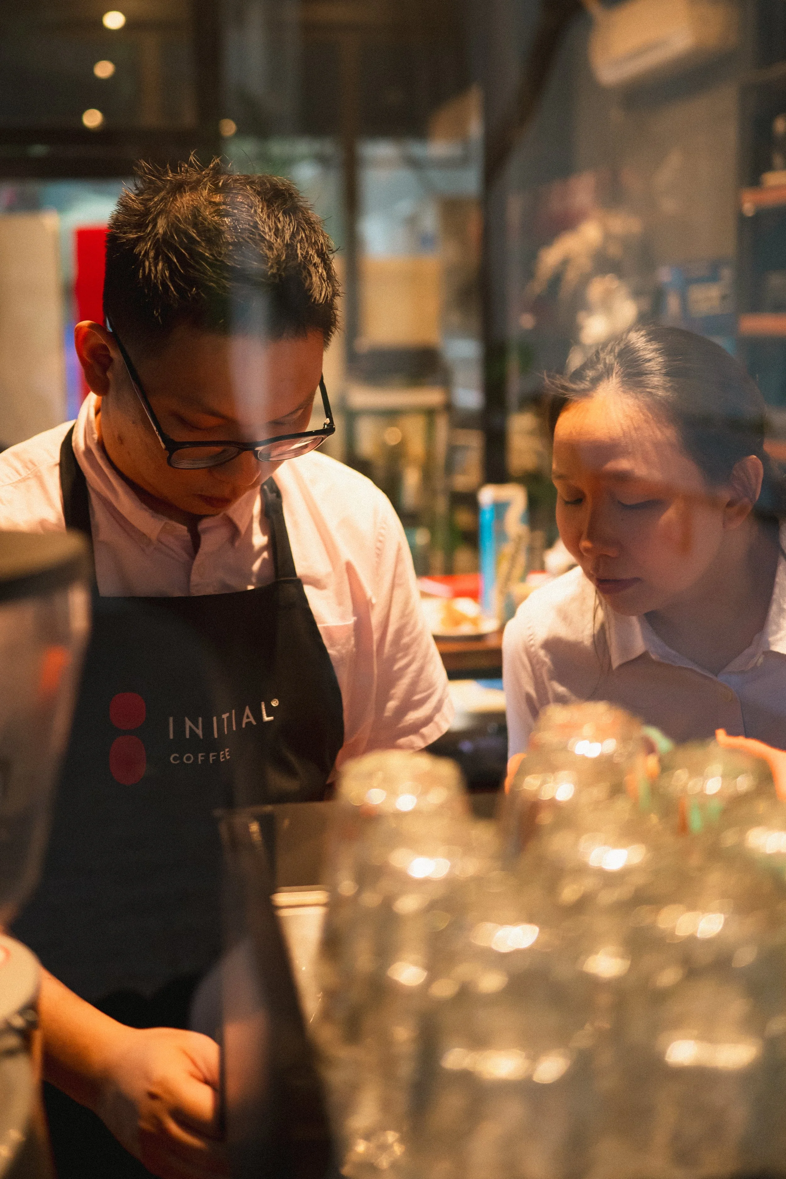 Two people working in a coffee shop, both wearing white shirts and black aprons. One person is wearing glasses and looking downward, while the other is looking to the side. The background is blurred with various items on shelves.