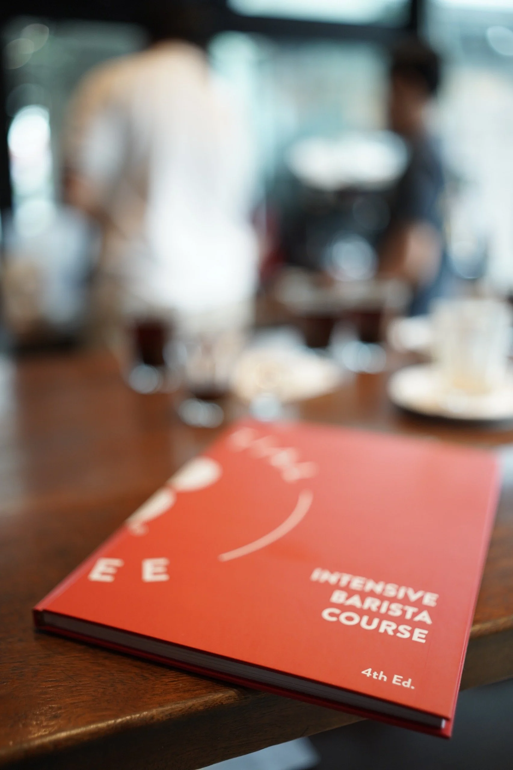 A red book titled 'Intensive Barista Course' resting on a wooden table. The book is 4th edition and the cover features white text and design elements.