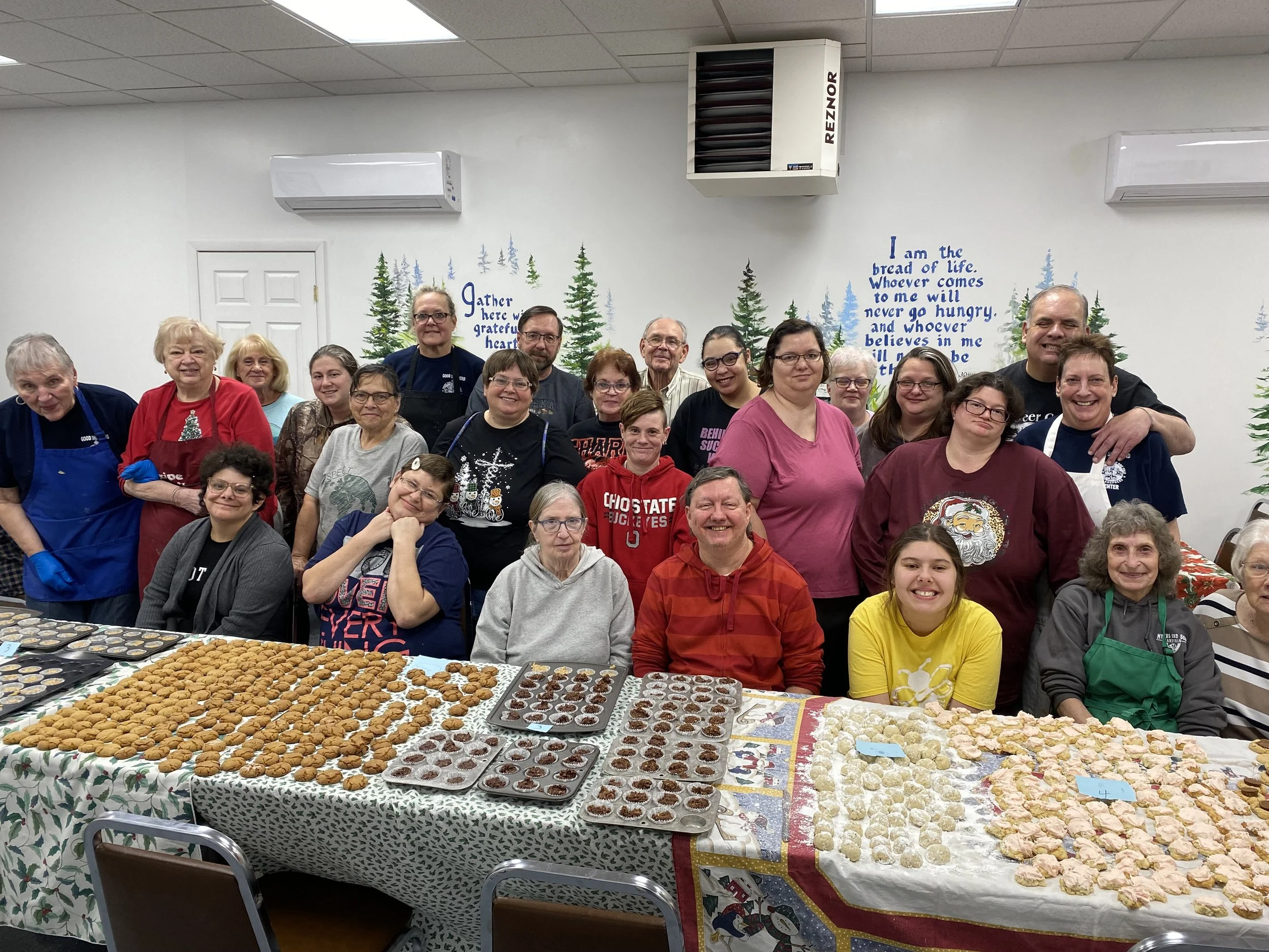 A group of people gathered in a festive kitchen, posing for a photo behind a long table filled with cookies and baked goods. The group includes adults and children, smiling and dressed in casual and holiday-themed clothing, with some wearing aprons. The background features a white wall decorated with trees and holiday quotes.