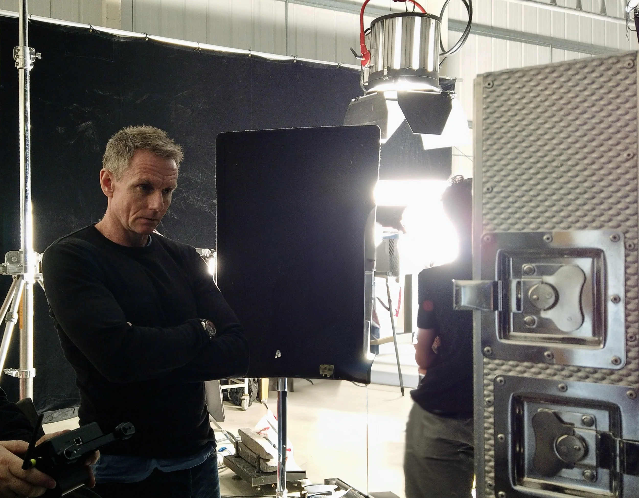 A man with short, light hair and a black shirt stands in a photography or filming studio, looking serious. The studio has equipment such as lights, soundproofing foam, and metallic frames, with black and white backgrounds.