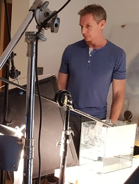 A man in a blue shirt looking at a glass box in a photography or filming setup, with lighting and camera equipment around.