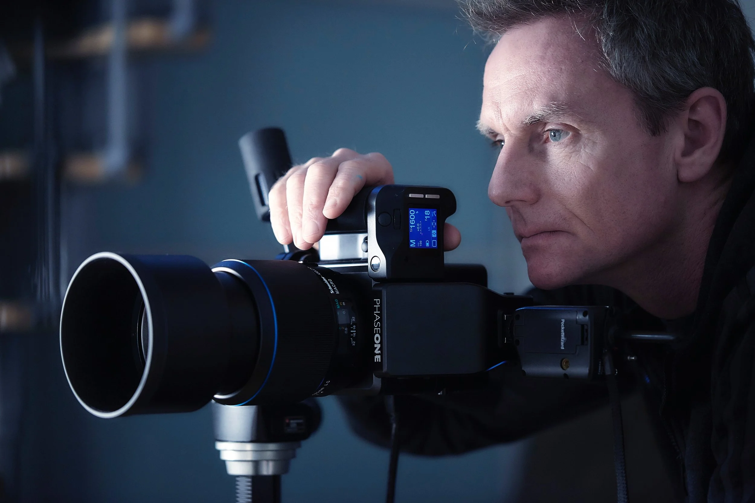 A man with blue eyes and dark hair looking through a large professional camera with a telephoto lens, in a dimly lit setting.