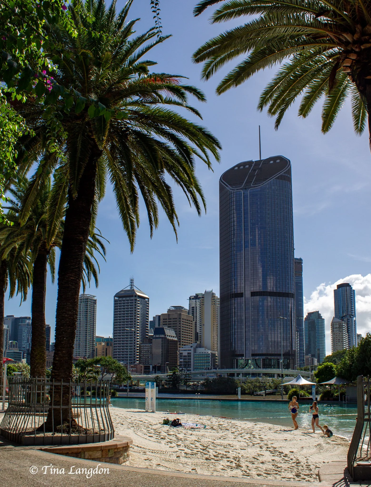 Swimmers coming out of the lagoon onto the sand at Streets Beach, South Bank, Brisbane, Australia surrounded by palm trees and skyscrapers.