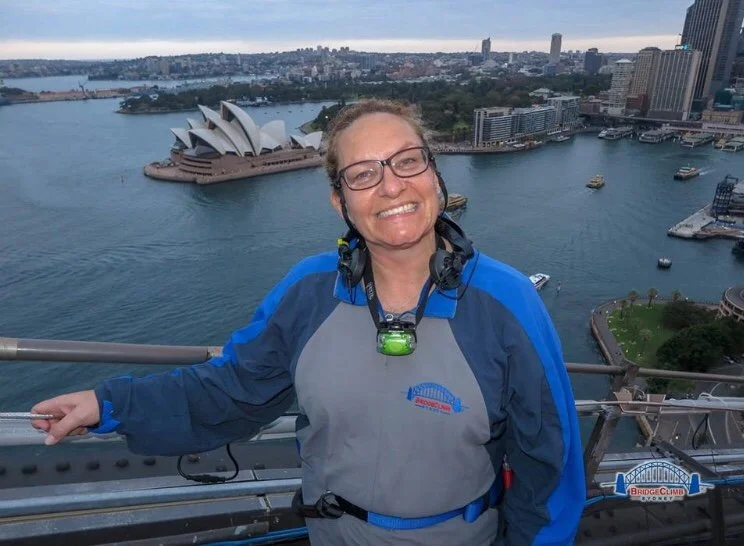 Smiling lady in blue and grey overalls standing on top of Sydney Bridge with the Opera House and harbour in the background