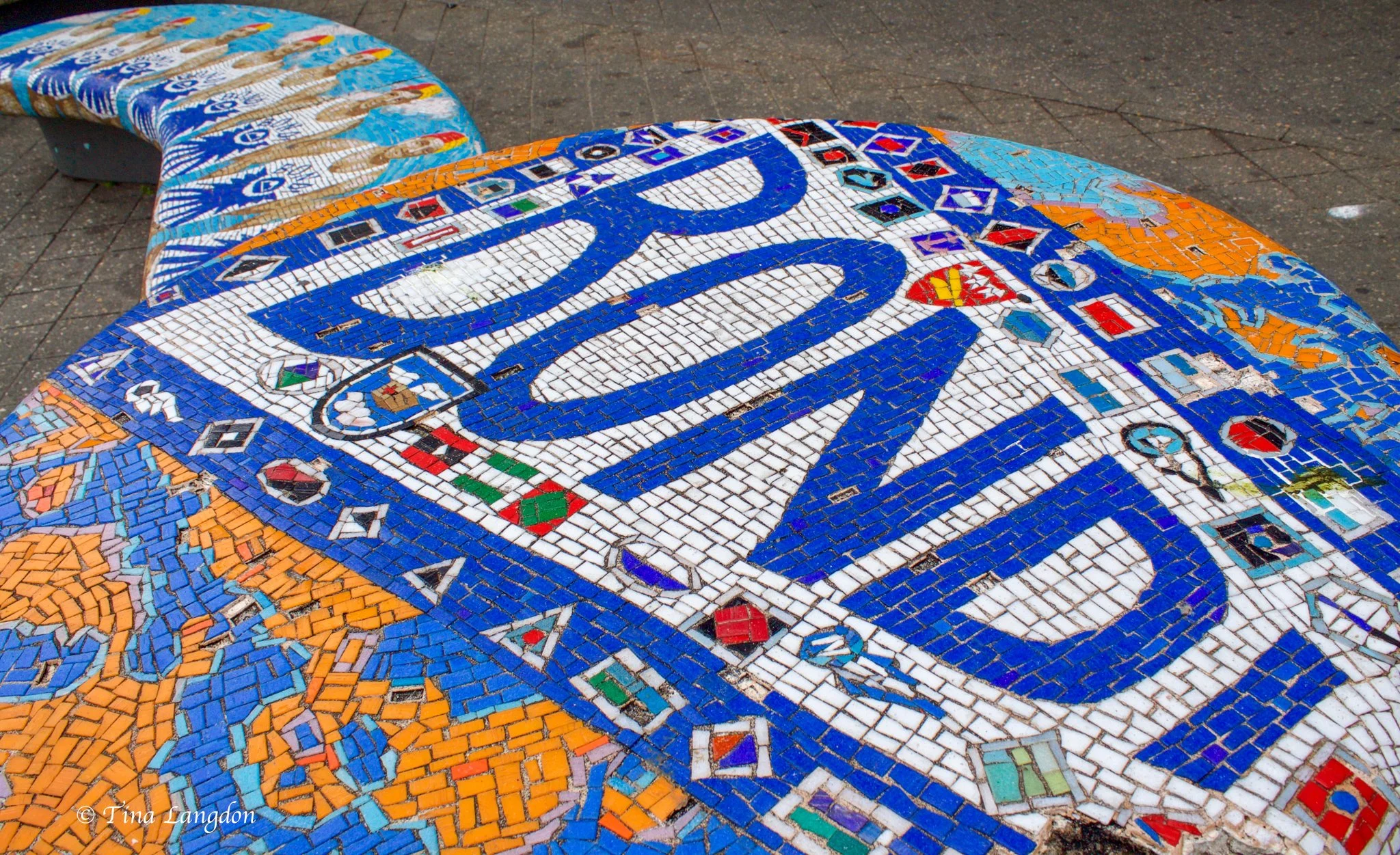 A brightly coloured mosaic beach seat with the letters for BONDI across the large round seat with seven sea swimmers standing  along the outstretched part of the seat