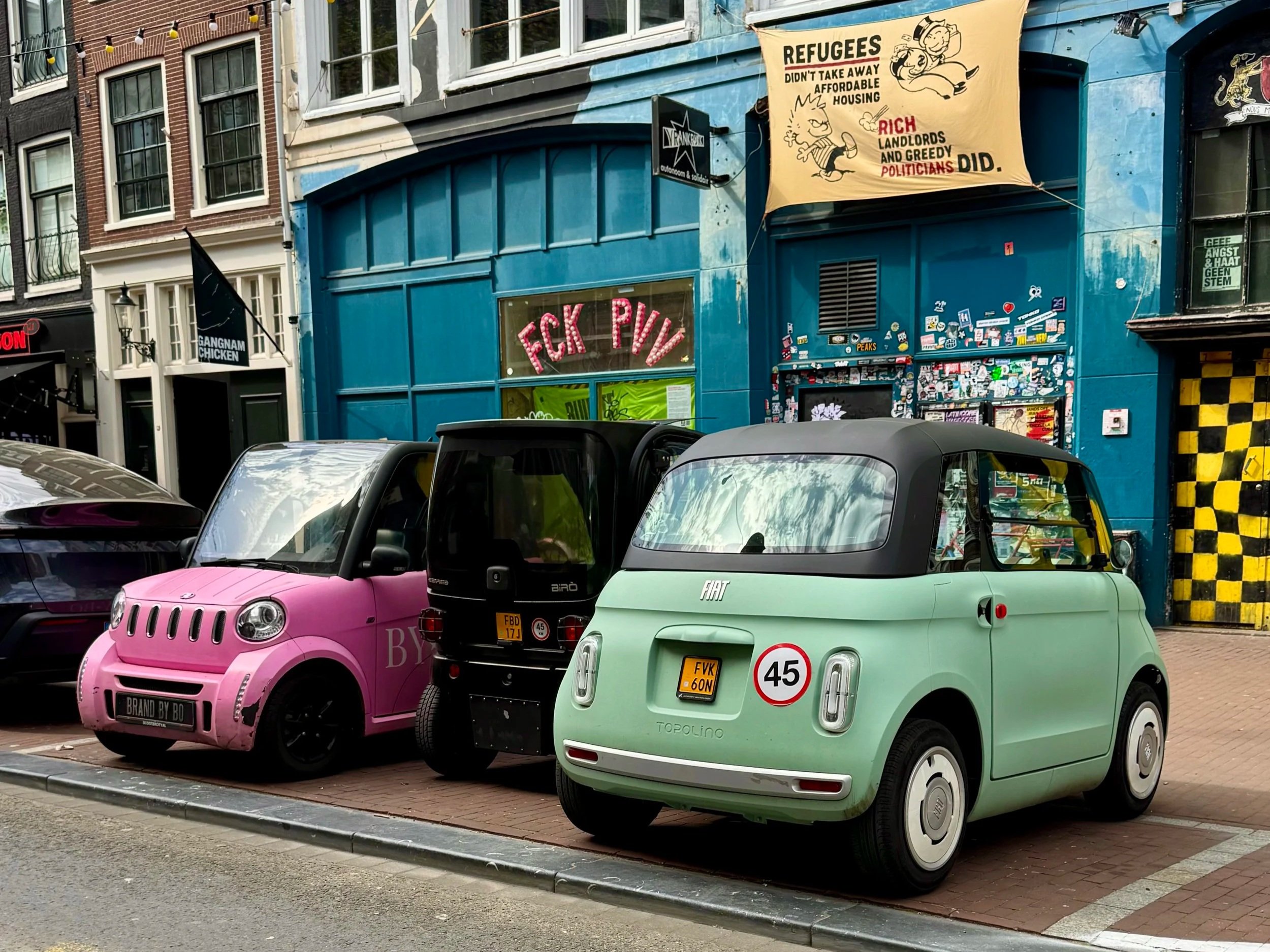Three tiny city cars, pink, black and green, parked next to each other in the space one standard car would take.