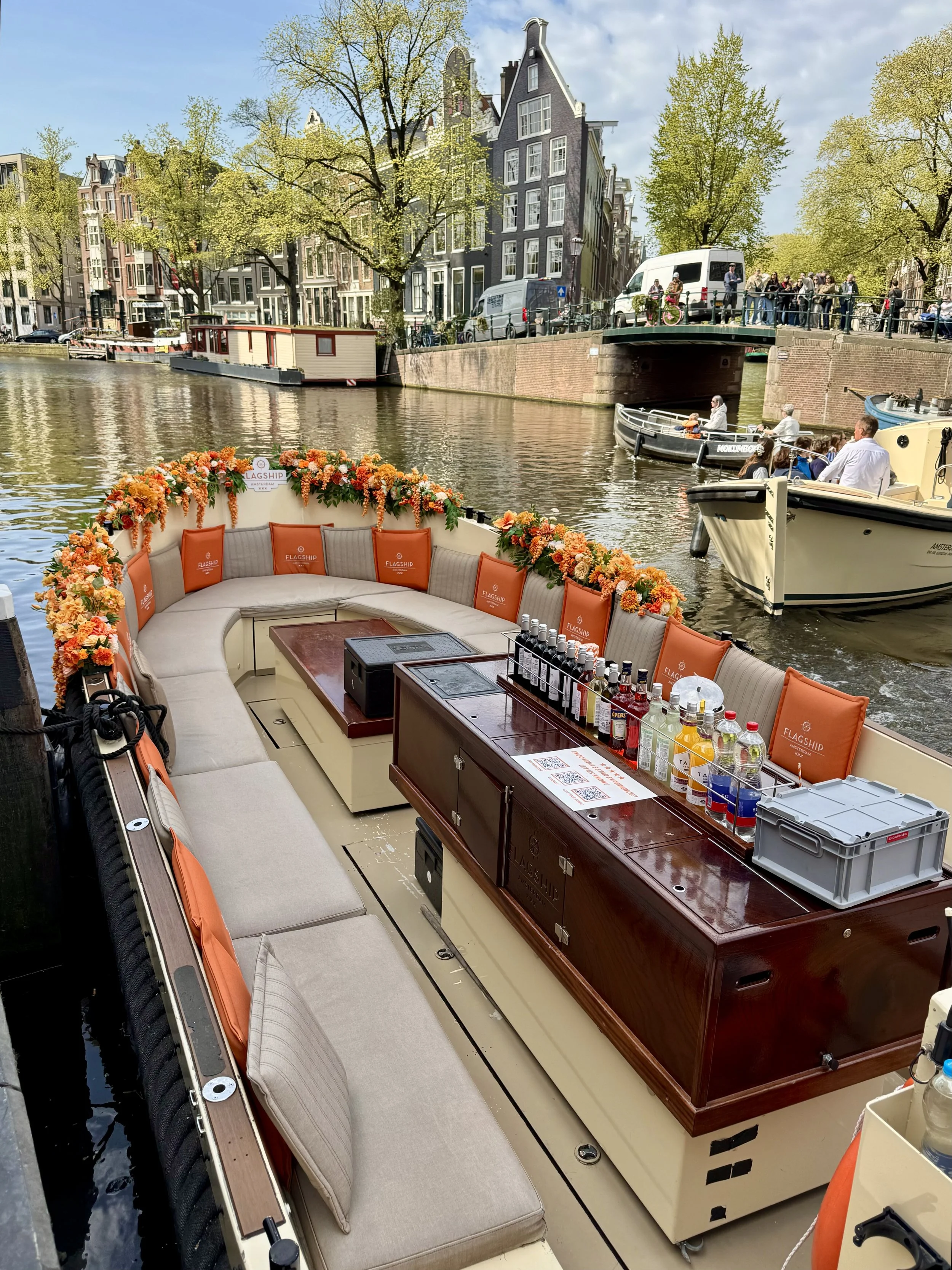 An empty canal boat decorated with orange flowers and alternating beige and orange cushions with a row of drinks lined up on a central wooden table takes up most of the photo. Two boats on the right by a low bridge. Tall thin buildings line the canal