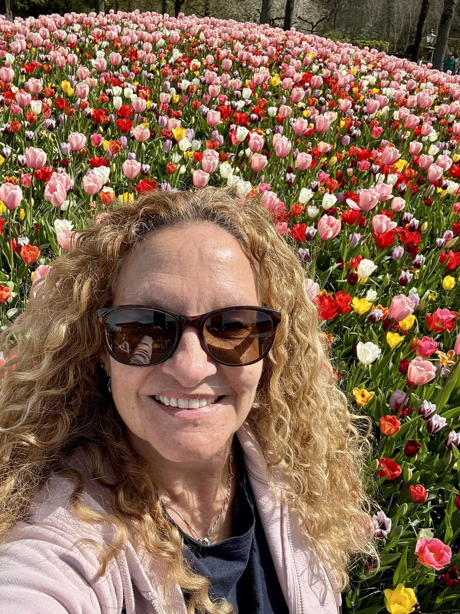 A smiling woman with long curly blond hair wearing sunglasses is taking a selfie while stood in front of a huge mound of brightly coloured tulips. The red, pink, white, orange, and yellow tulips are in full bloom.