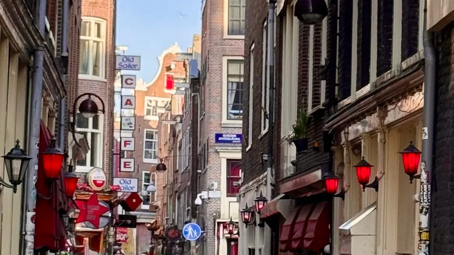 Small red street wall lights above doorways line a narrow lane of brown brick buildings. A vertical street sign spelling CAFE is in the background.