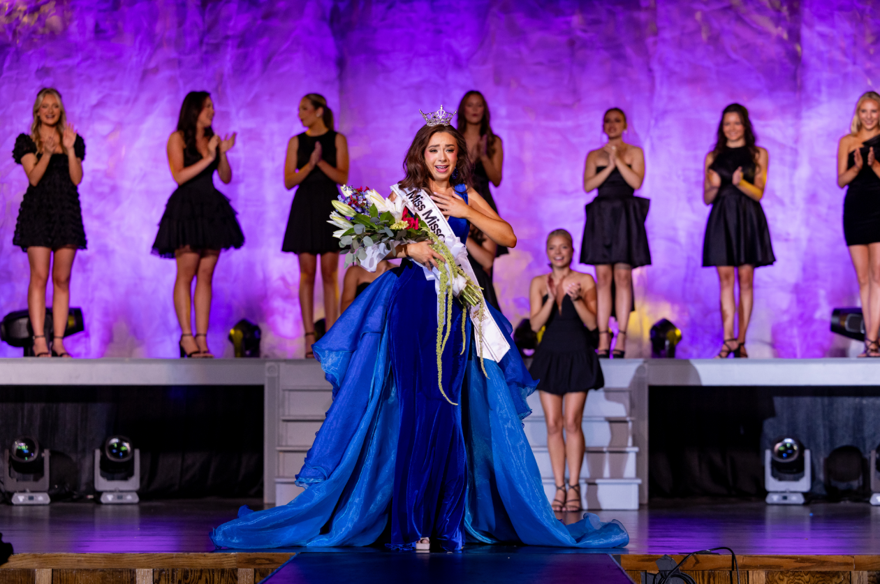Color photo of a beauty pageant winner receiving a crown. A woman with a crown is being crowned by another woman. The crowned woman appears joyful and emotional, wearing a sash and statement earrings.