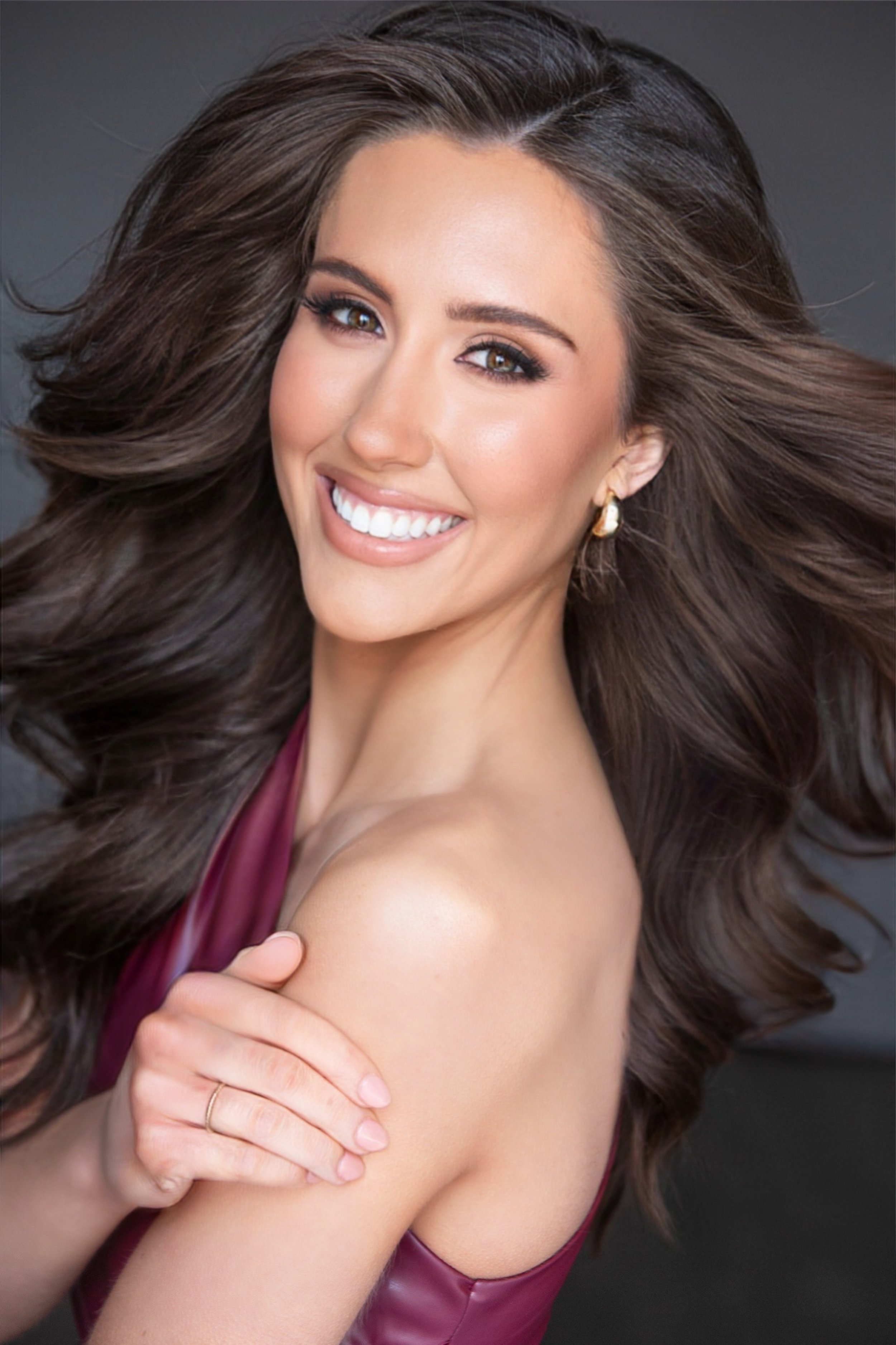 Smiling woman with long dark hair, wearing a purple blouse and sparkly earrings. She is posing against a neutral background.