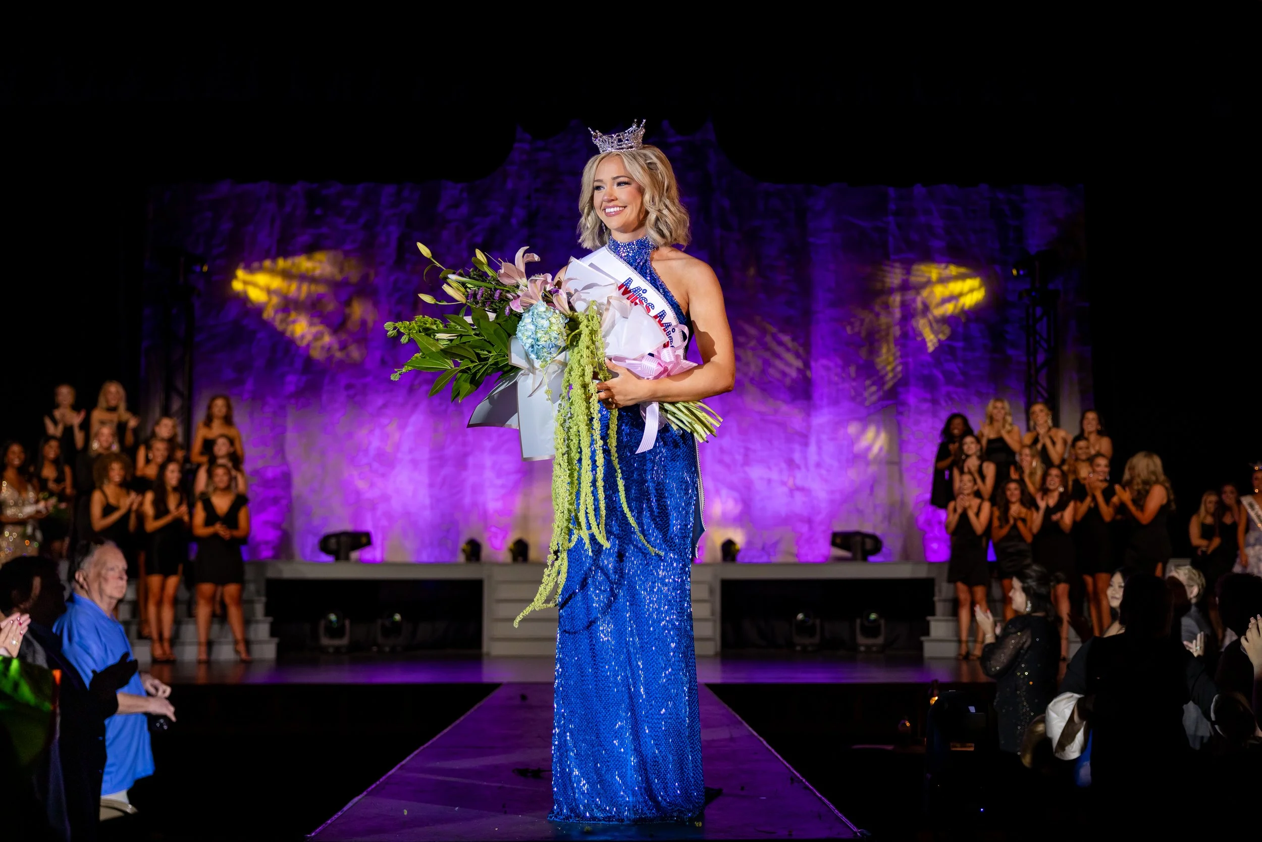 Black and white photo of a pageant winner wearing a crown and holding a bouquet of flowers, with a sash over her shoulder. She is on stage with a spotlight shining on her.