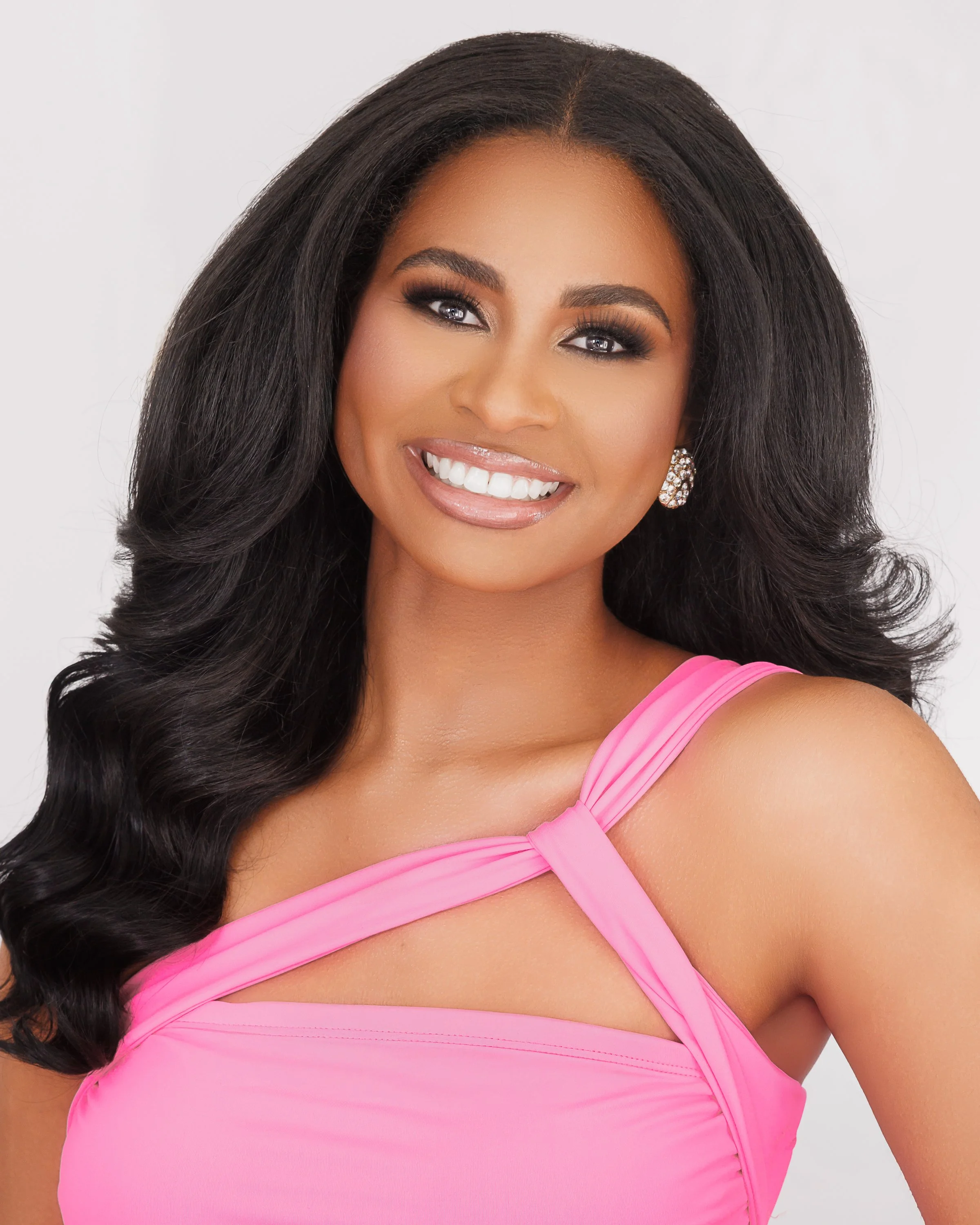 Woman smiling in purple dress with flower detail on shoulder standing outdoors.