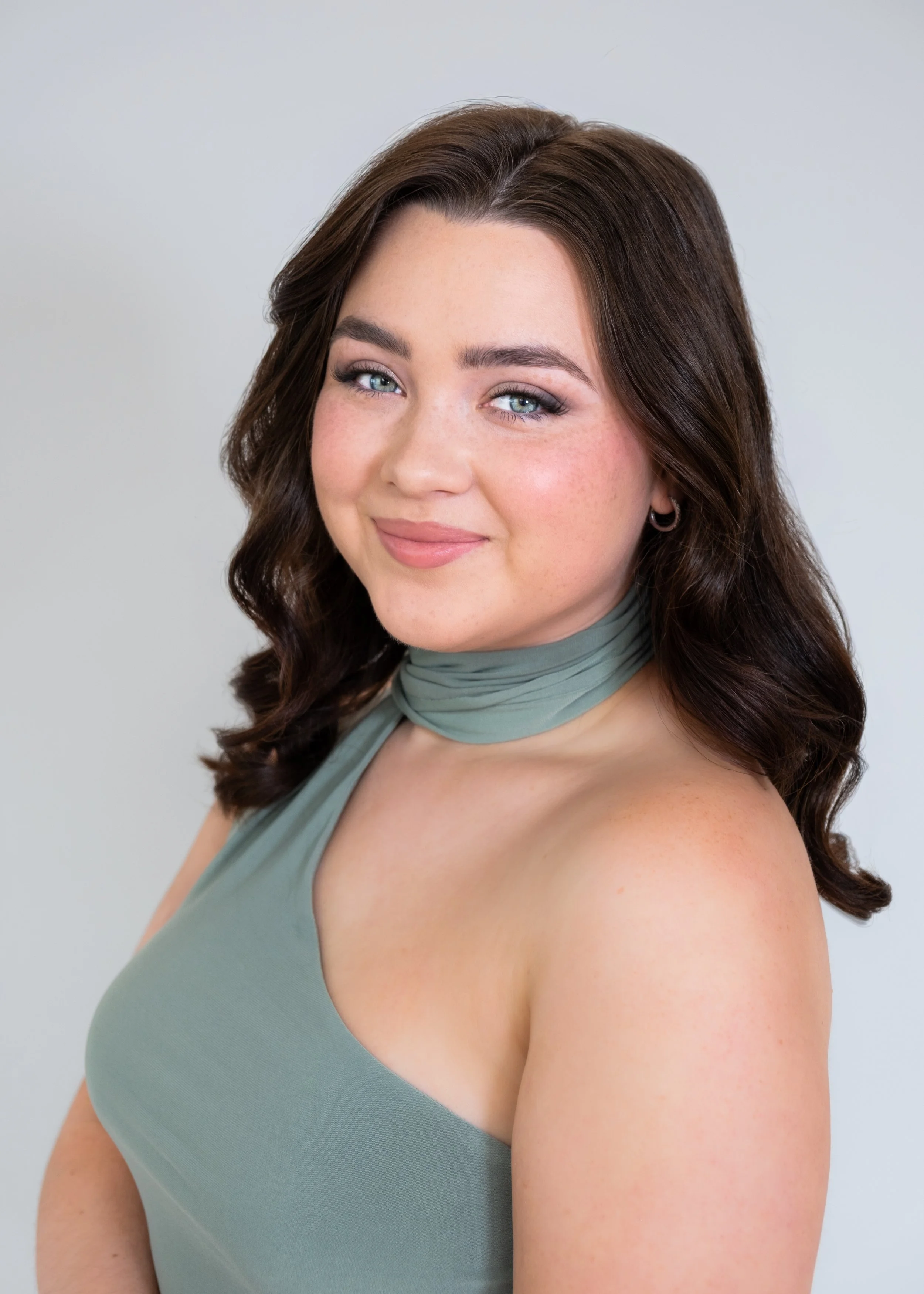 Portrait of a woman with long black hair and red lipstick, wearing a strapless top, leaning her face on her hand against a neutral backdrop.
