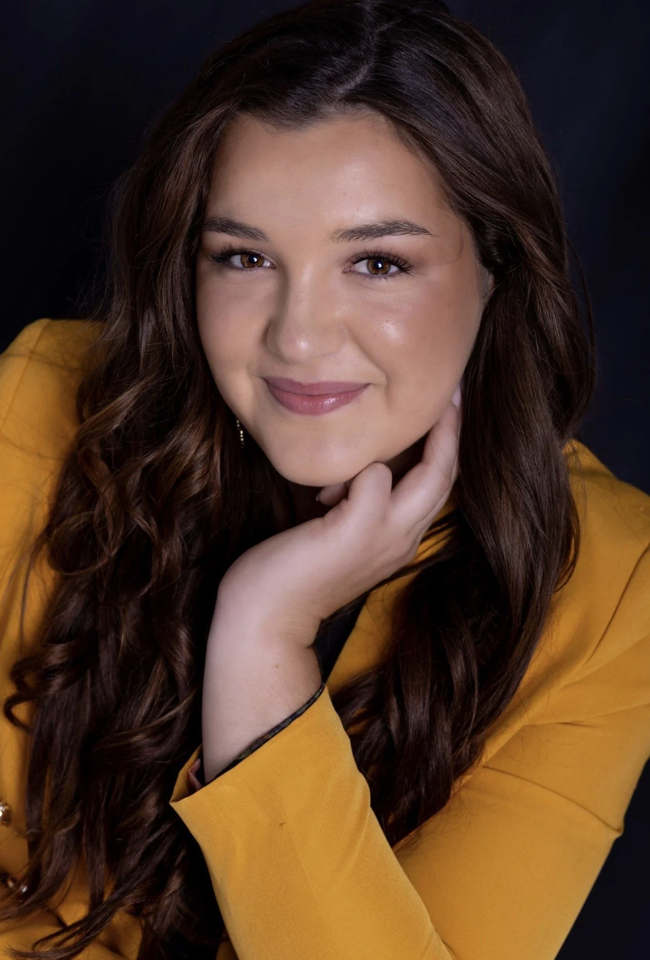Portrait of a woman with long black hair and red lipstick, wearing a strapless top, leaning her face on her hand against a neutral backdrop.