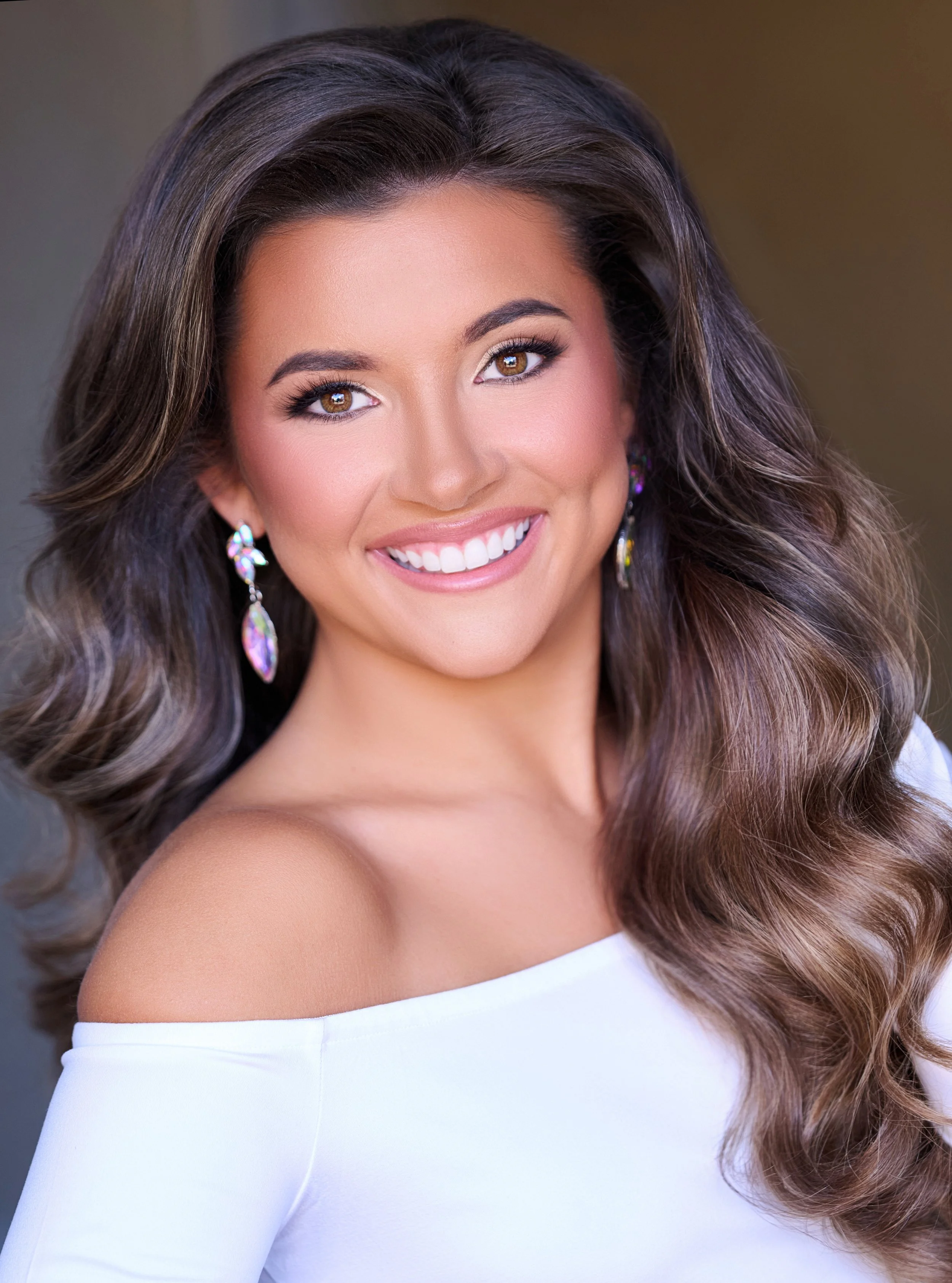 Woman with long blonde hair wearing a pink top and large, decorative earrings, posing against a dark background.