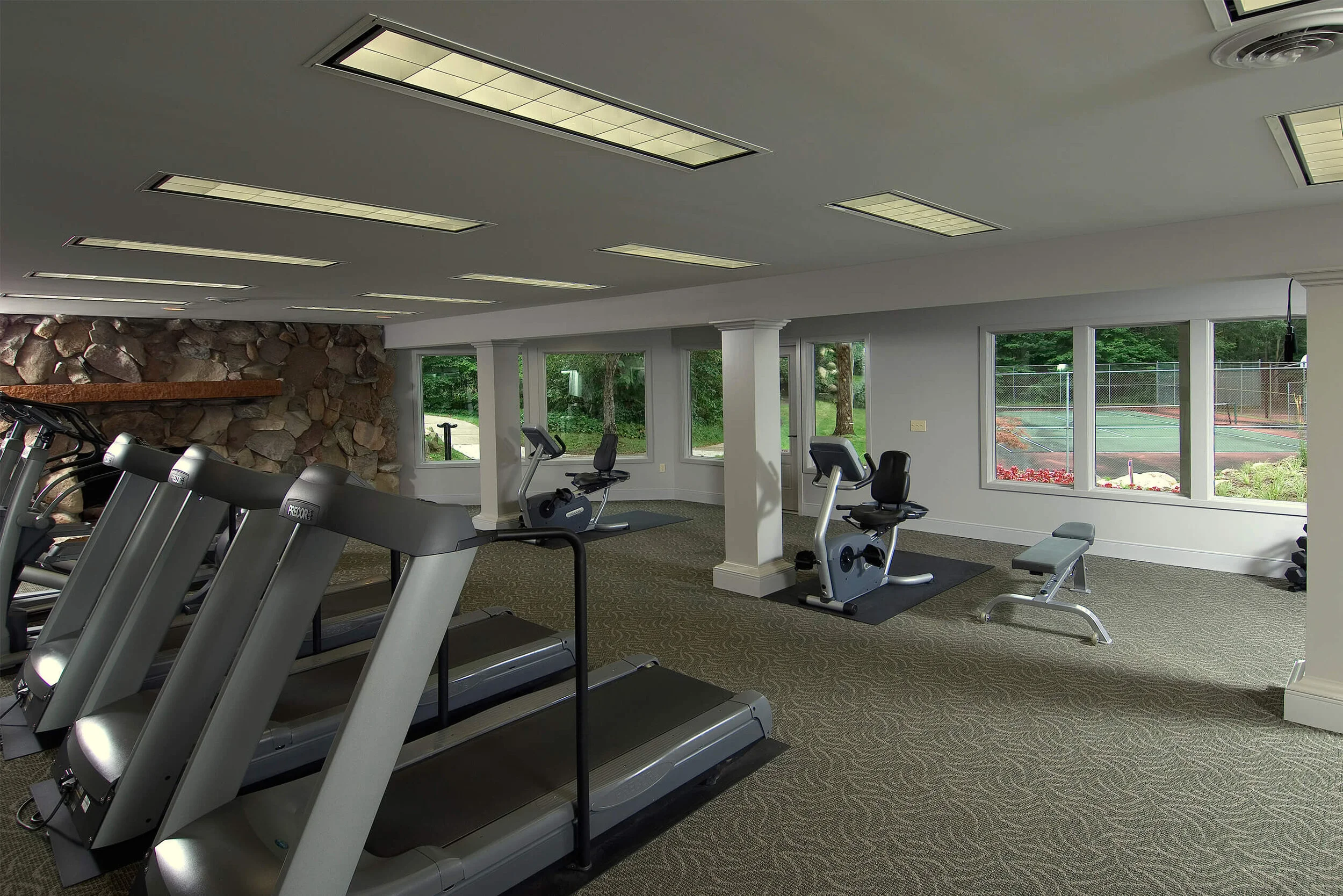 Interior photo of apartment club house gym with treadmills, exercise bike and weight bench. The gym has large windows that include a view of the exterior tennis court. 