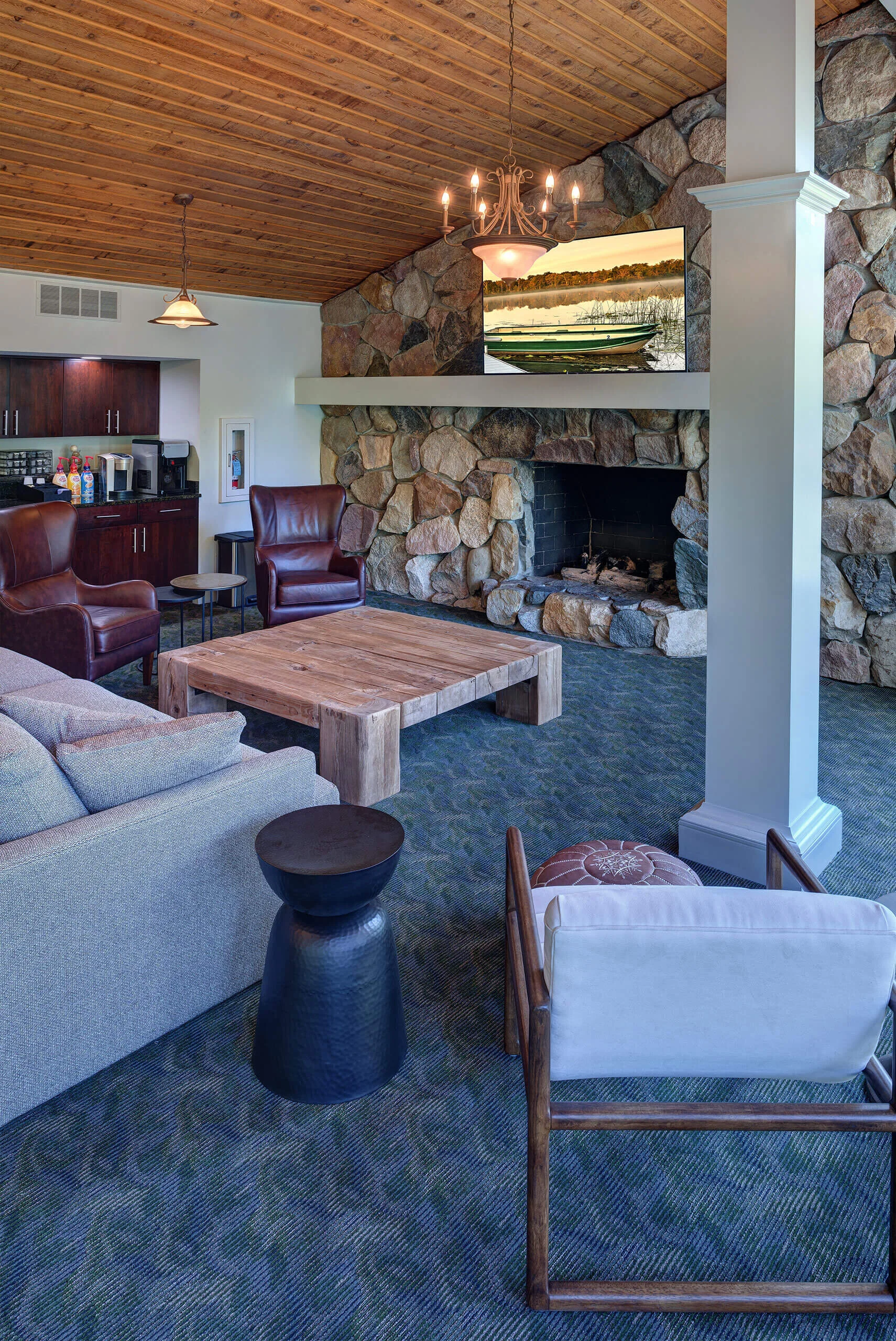 Interior photo of apartment clubhouse with couch, leather chairs and a kitchen. The room has high wood ceilings, a large stone fireplace and a carpeted floor. 