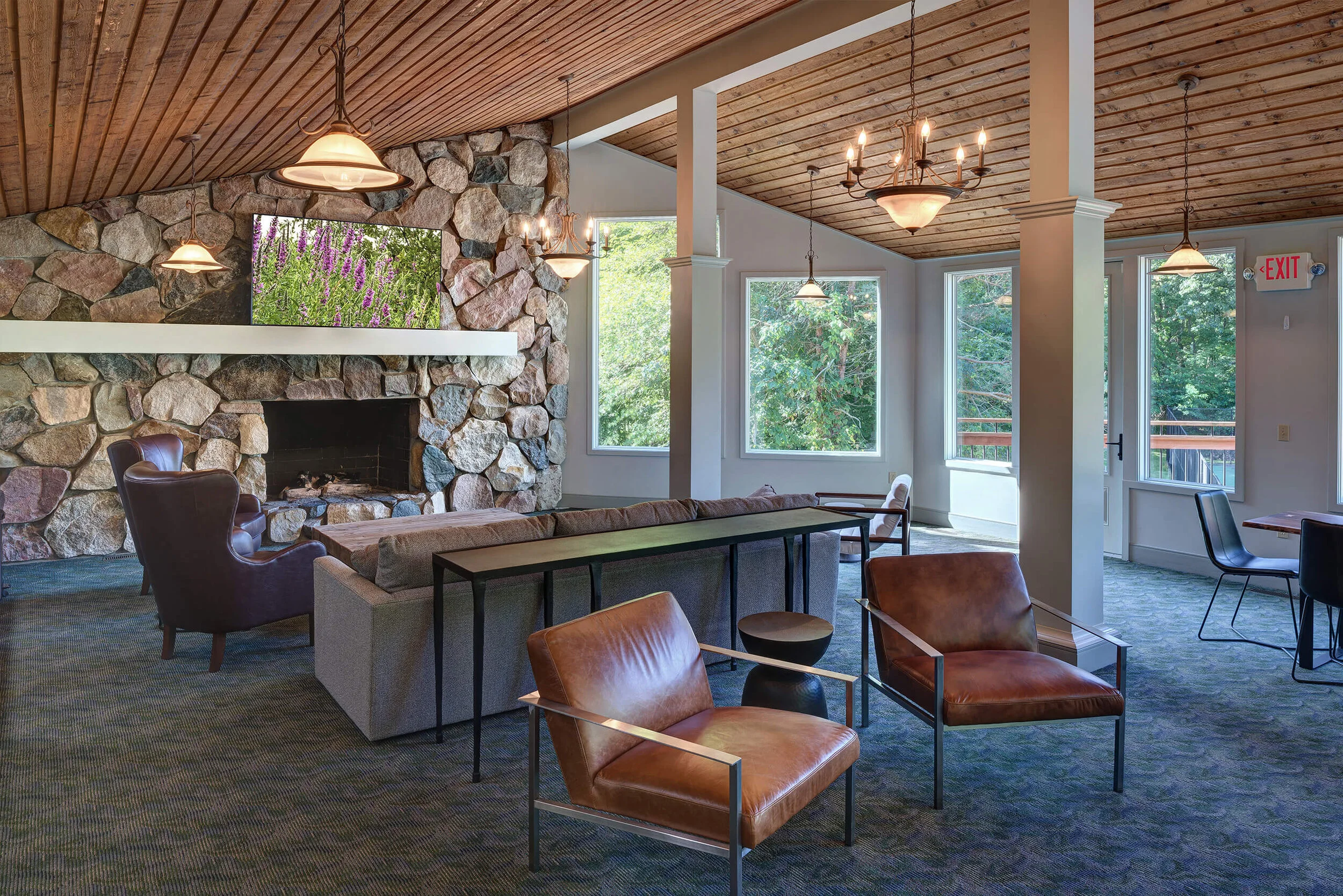 Interior photo of apartment clubhouse with couch, leather chairs and a kitchen. The room has high wood ceilings, a large stone fireplace and a carpeted floor. 