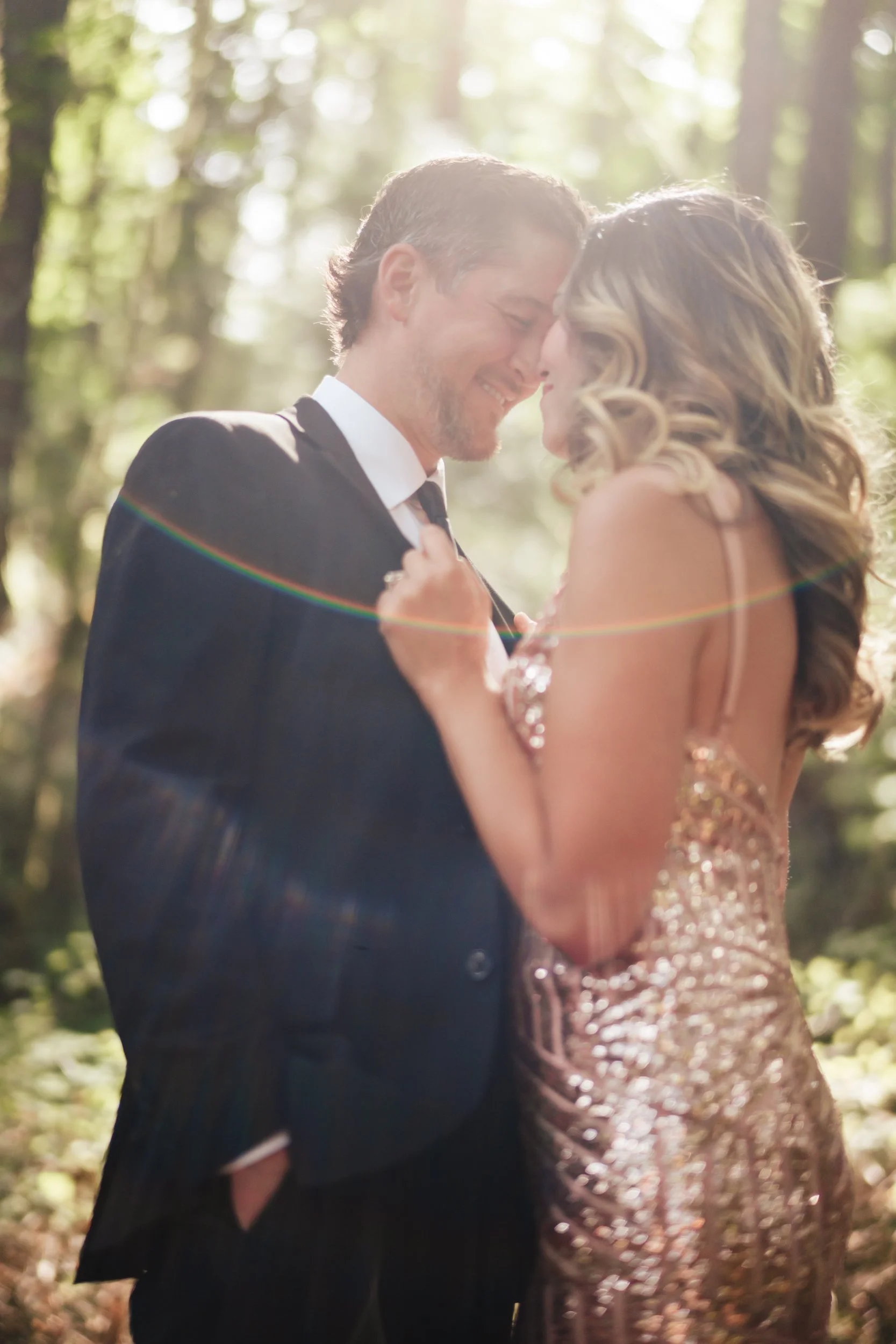 A woman wearing a shimmery dress pulls her husband closer while a rainbow sunburst illuminates the scene.