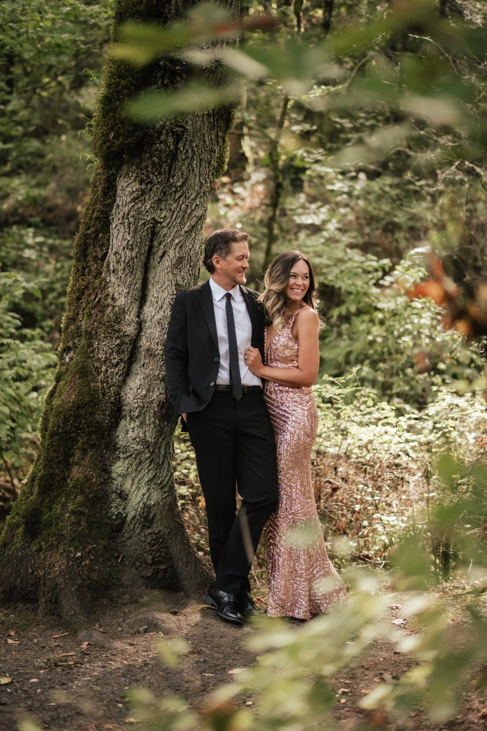 A man wearing a black tie and suit leans against an old tree with his wife in Forest Park.