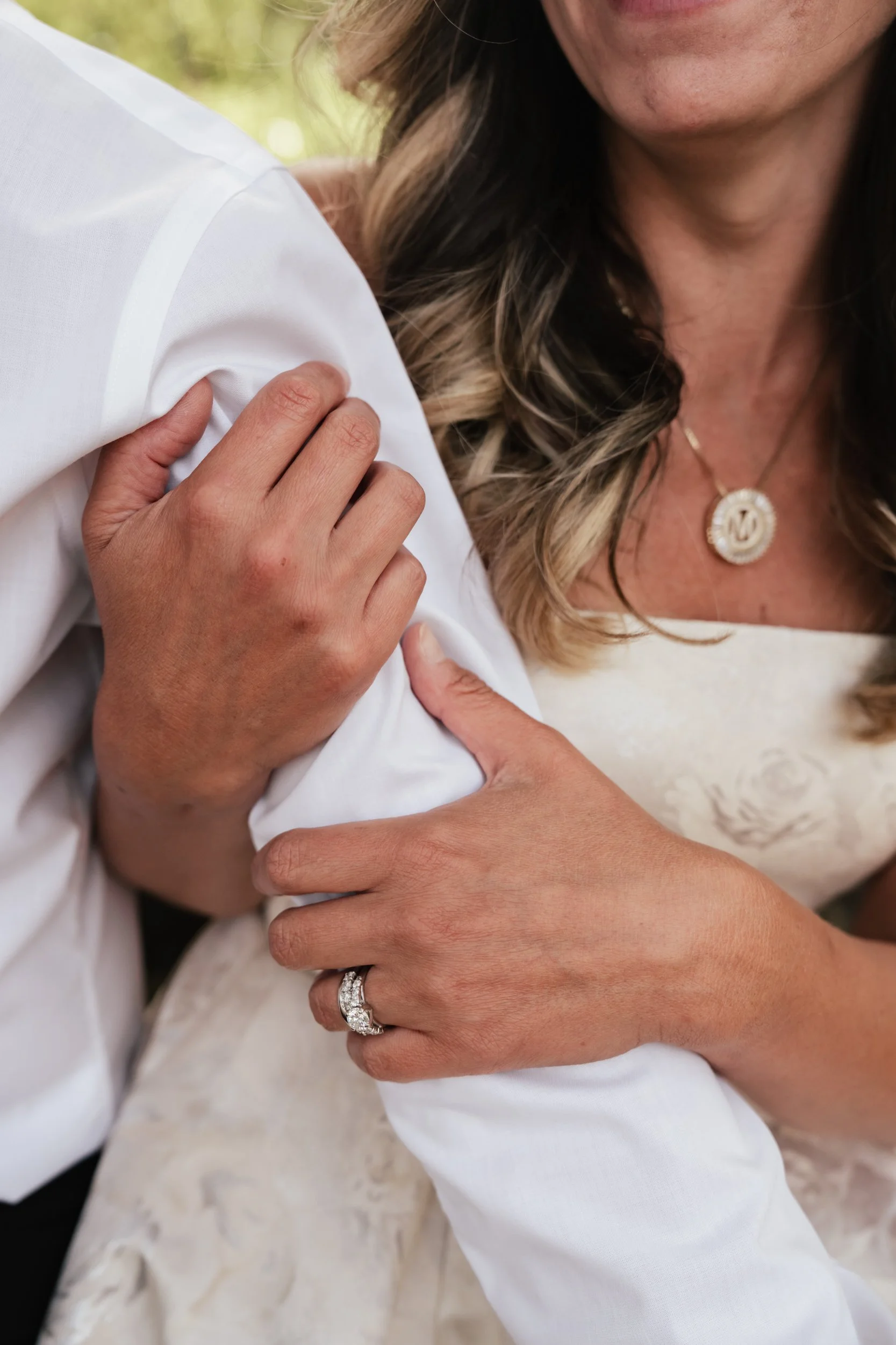 A woman with an engagement ring and diamond necklace squeezes the arm of her man wearing a white button down shirt.