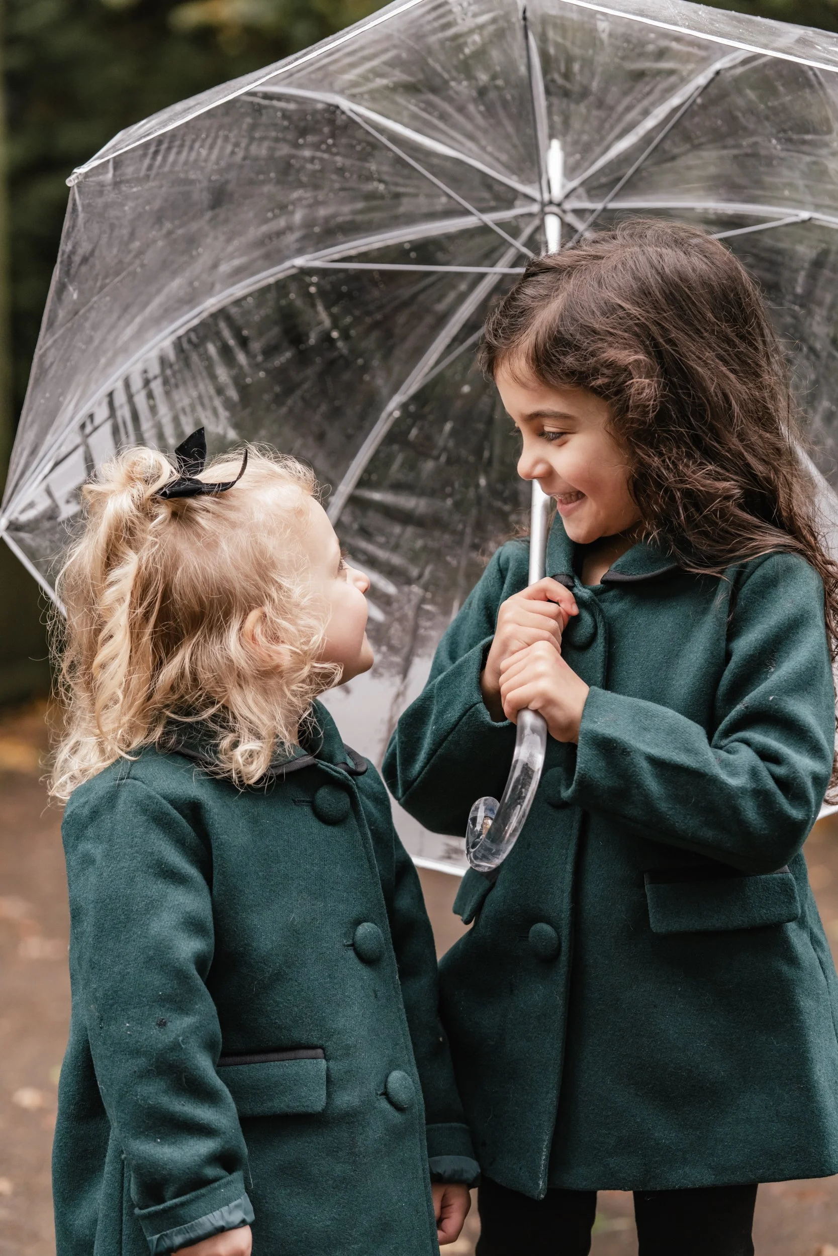Two sisters in deep green coats share a clear umbrella.