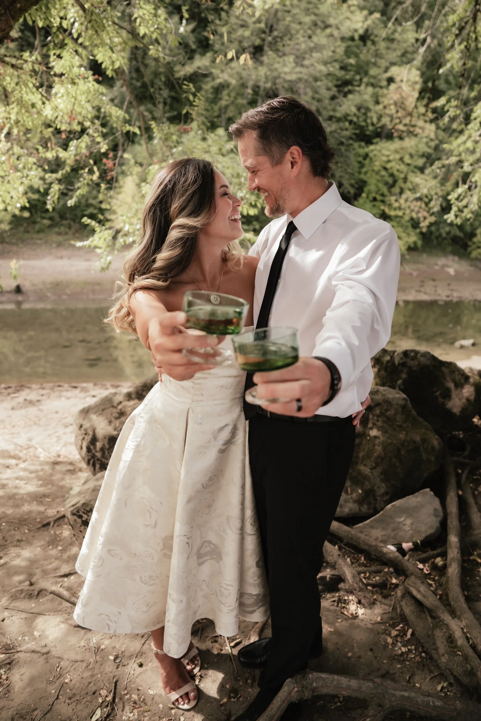 A newly engaged couple cheers to their future together along a rocky, tree lined river.